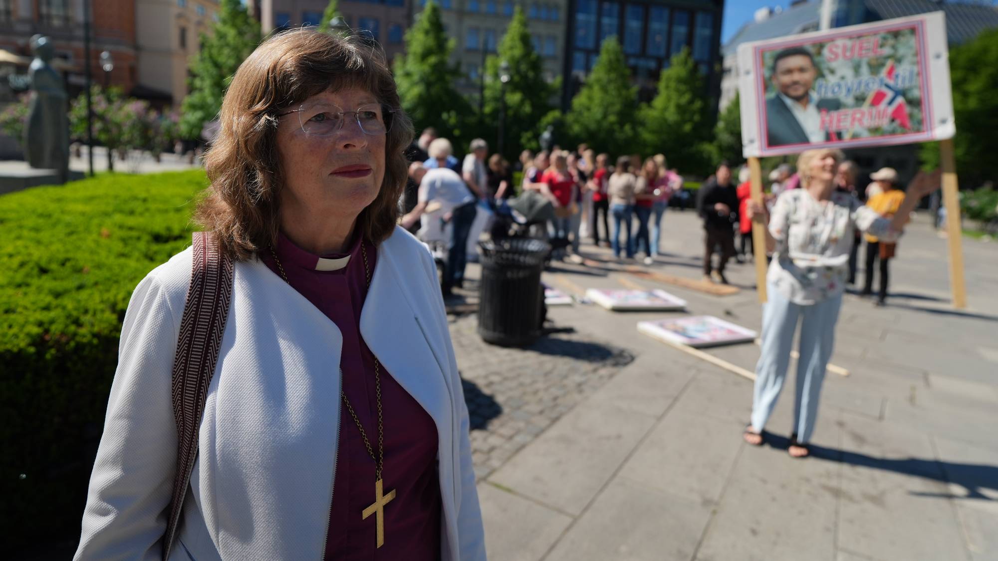 Ingeborg Middtømme har på seg kvit jakke og prestekrage. Ho står framfor Stortinget, og i bakgrunnen kan ein sjå demonstrantar. 