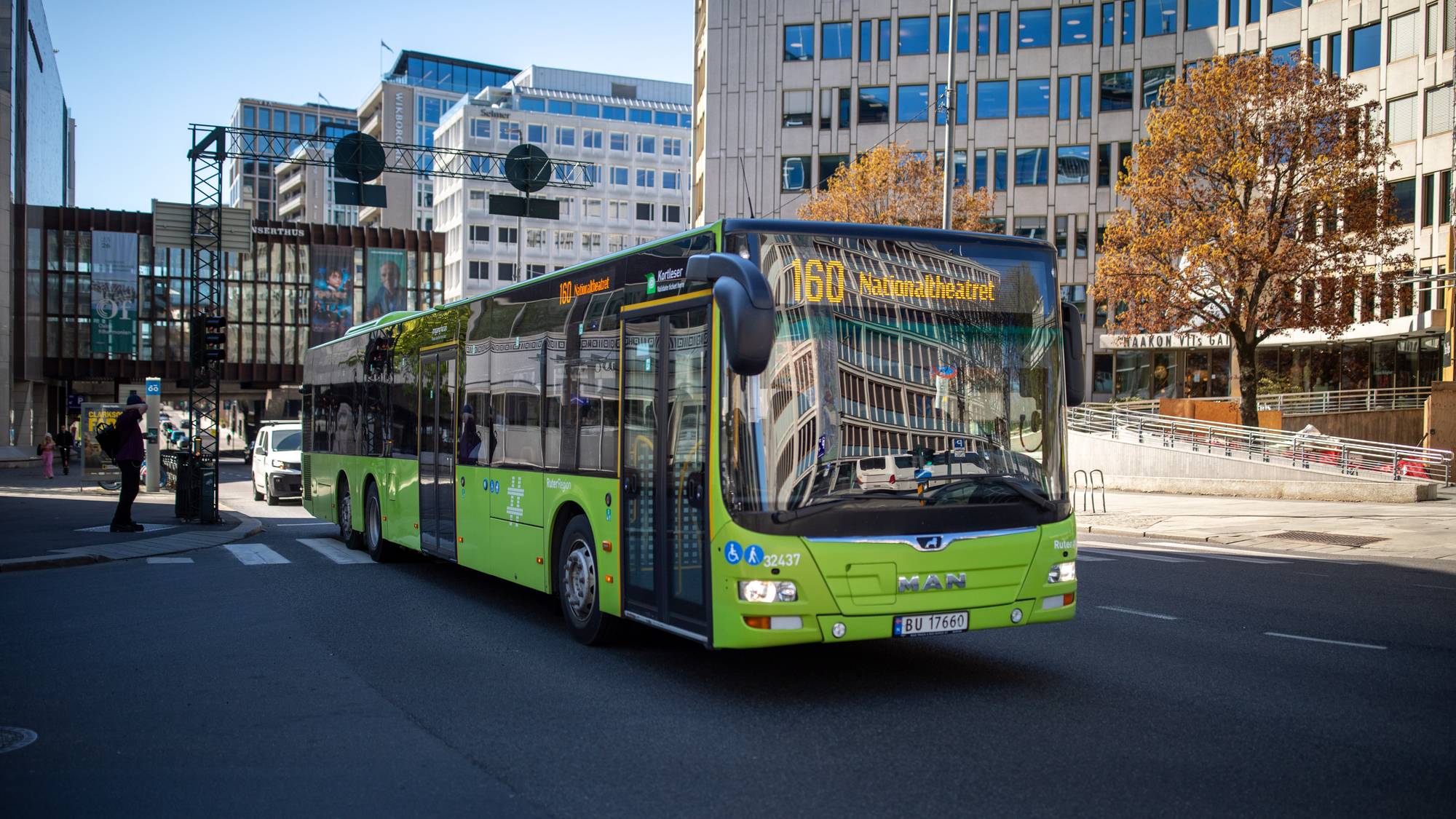 En grønn MAN-buss kjører på en gate i sentrum av Oslo. Bussen har rutenummer 160 og destinasjonen Nationaltheatret. I bakgrunnen sees flere bygninger og et tre med gulnede blader. (Bildebeskrivelsen er laget av en KI-tjeneste)