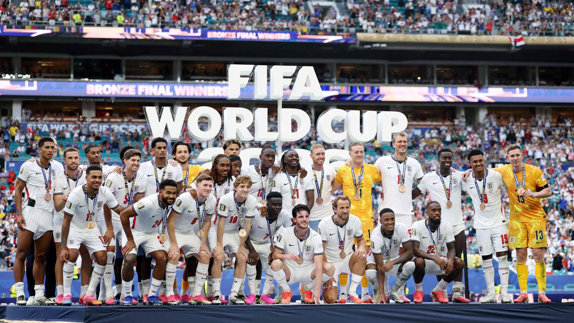 Det engelske landslaget i fotball poserer med bronsemedaljer foran en FIFA-logo på et fullsatt stadion. (Bildebeskrivelsen er laget av en KI-tjeneste)