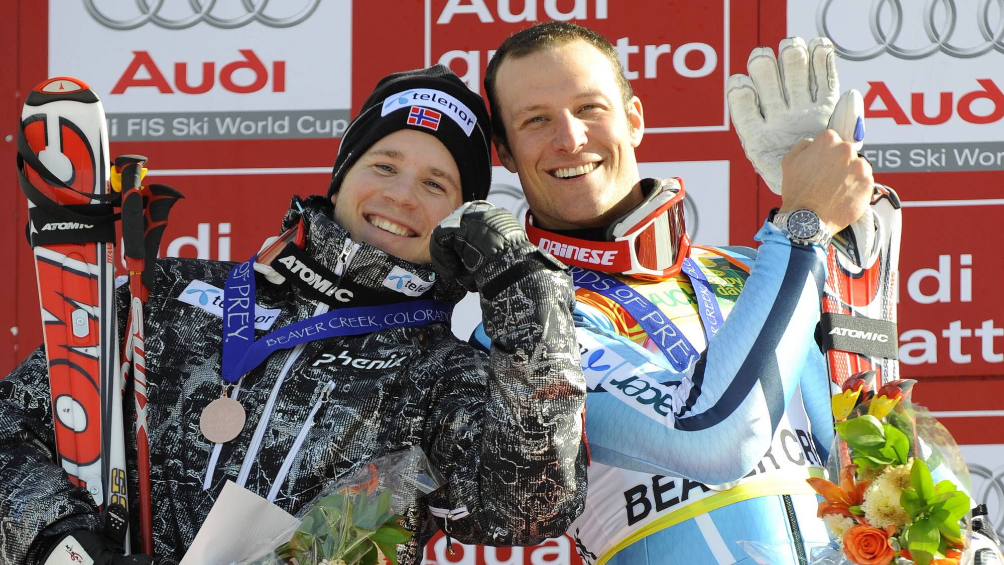 To skiløpere står på et podium og smiler. Den ene holder fram en bronsemedalje. Begge har skistaver og ski. Bak dem vises et rødt banner med Audi-logo. (Bildebeskrivelsen er laget av en KI-tjeneste)