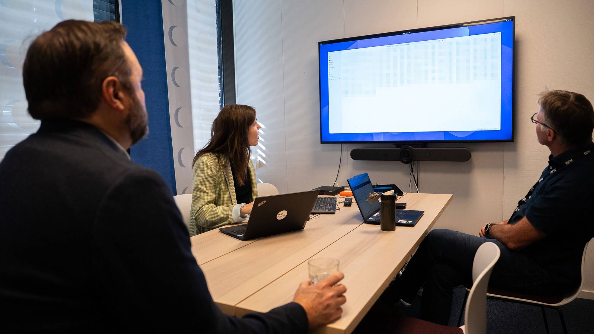 To menn og en dame kikker vendt mot en stor tv-skjerm, skjermen har filer som er blurret ut. 