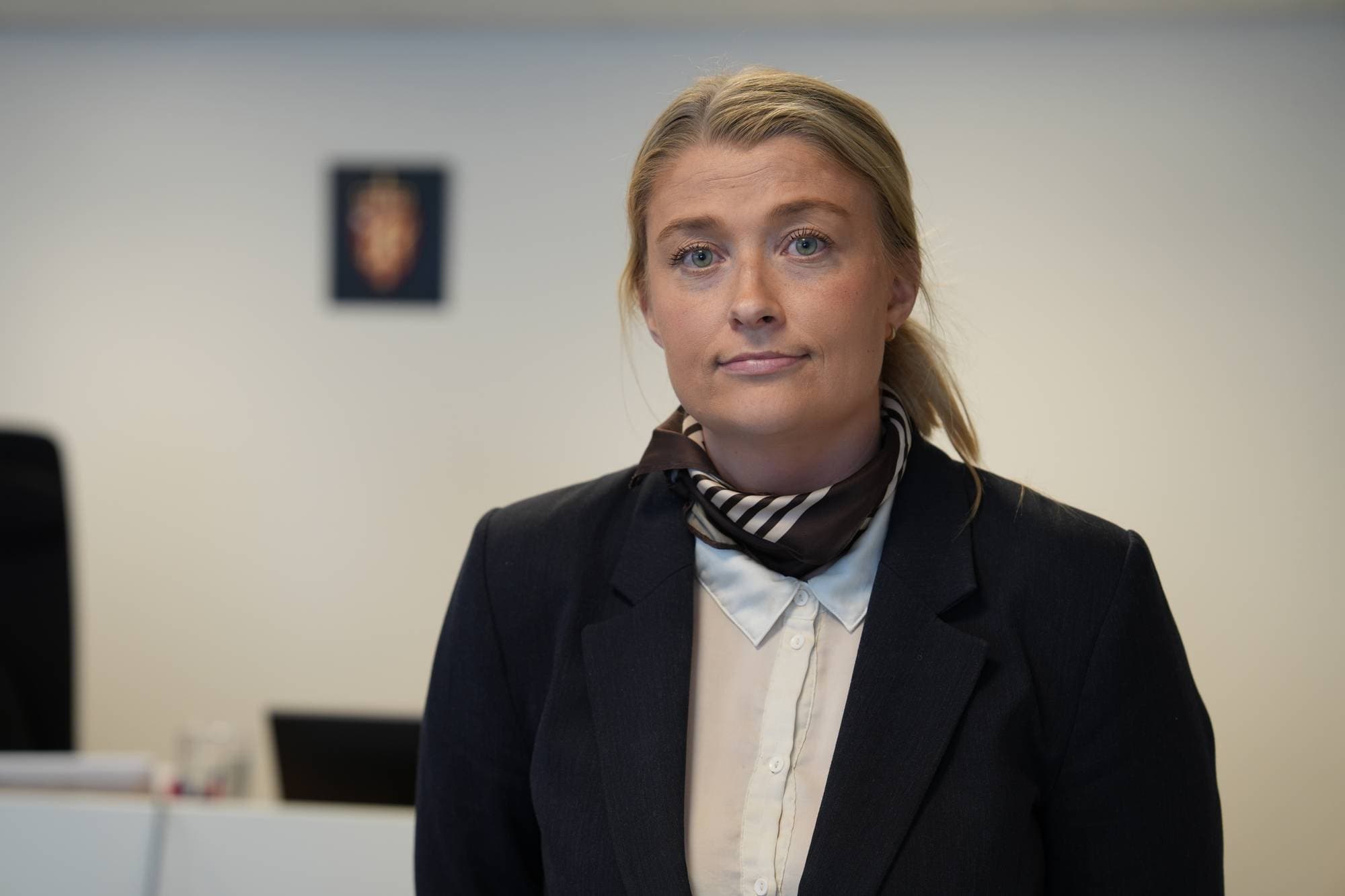 En kvinne med blondt hår, kledd i mørk blazer og silkeskjerf, ser mot kamera. Bak henne, på en lys vegg, henger et emblem. Rommet ser ut til å være et kontor. (Bildebeskrivelsen er laget av en KI-tjeneste)