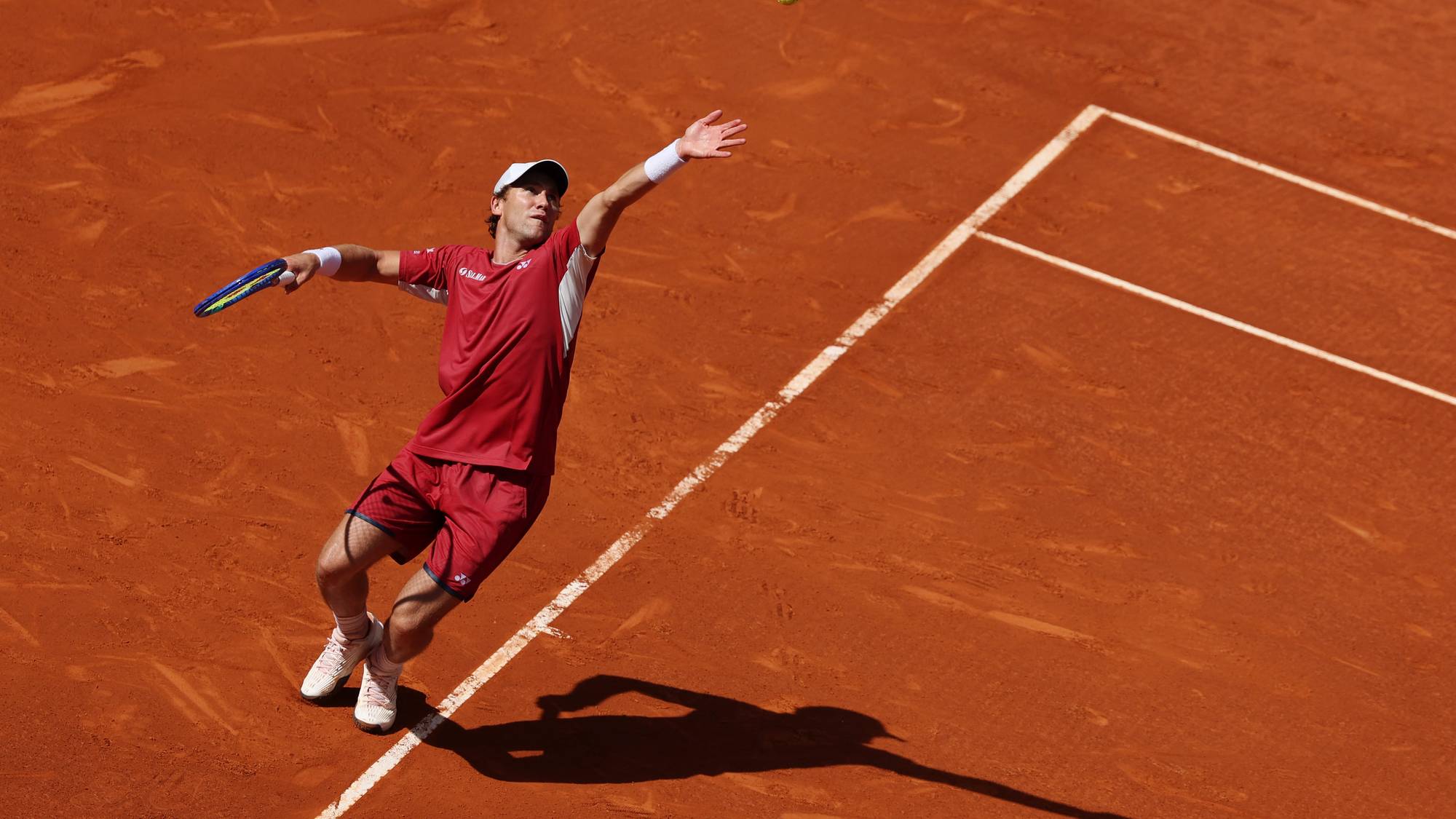 En tennisspiller i en rød overdel og shorts kaster opp en ball for å serve. Han står på en grusbane med hvite linjer. Et stort, hvitt NTB-symbol er plassert midt på bildet. (Bildebeskrivelsen er laget av en KI-tjeneste)