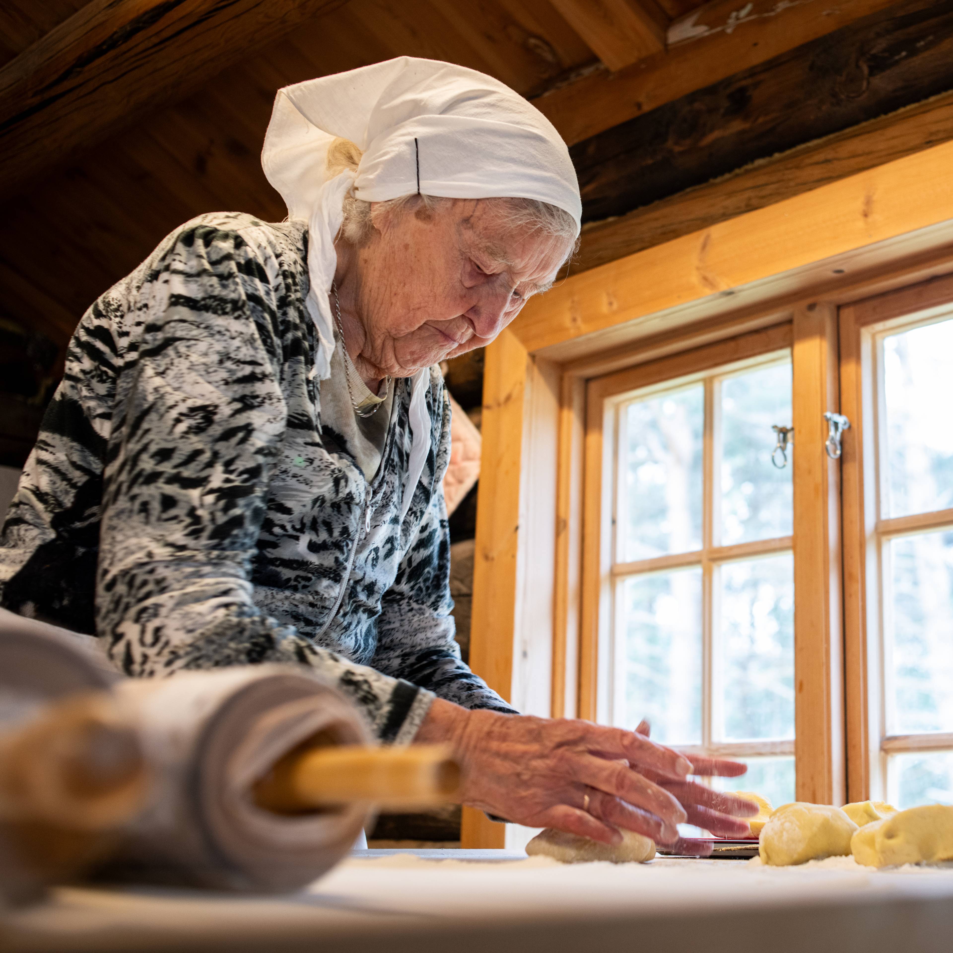 Eldre kvinne med hodeplagg baker med deig i et tømmerhus ved et vindu.