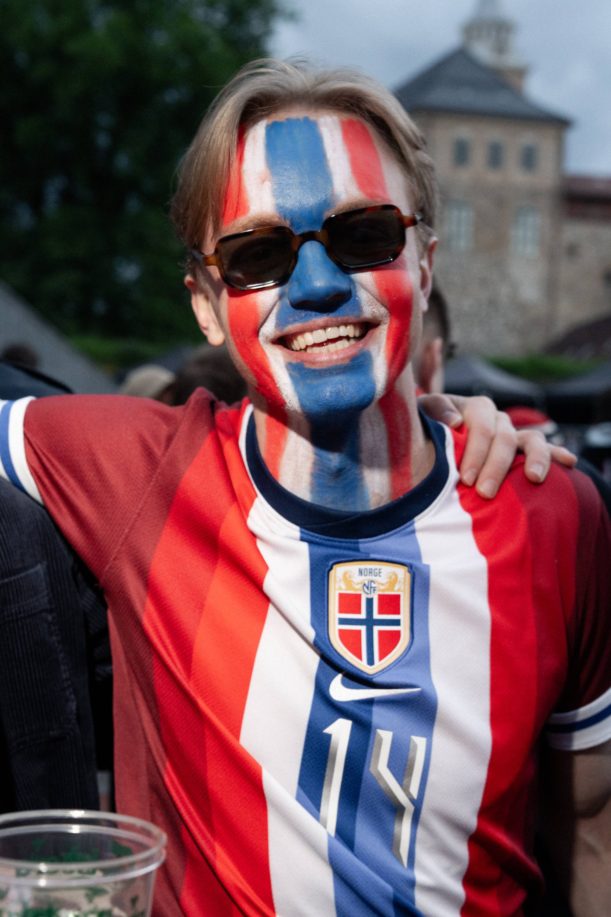 En mann med norsk flagg malt i ansiktet og på halsen smiler til kameraet.
Han har på seg en norsk landslagsdrakt.
I bakgrunnen er det mennesker og en bygning som ligner et slott.
Bildet er tatt utendørs i dagslys. (Bildebeskrivelsen er laget av en KI-tjeneste)