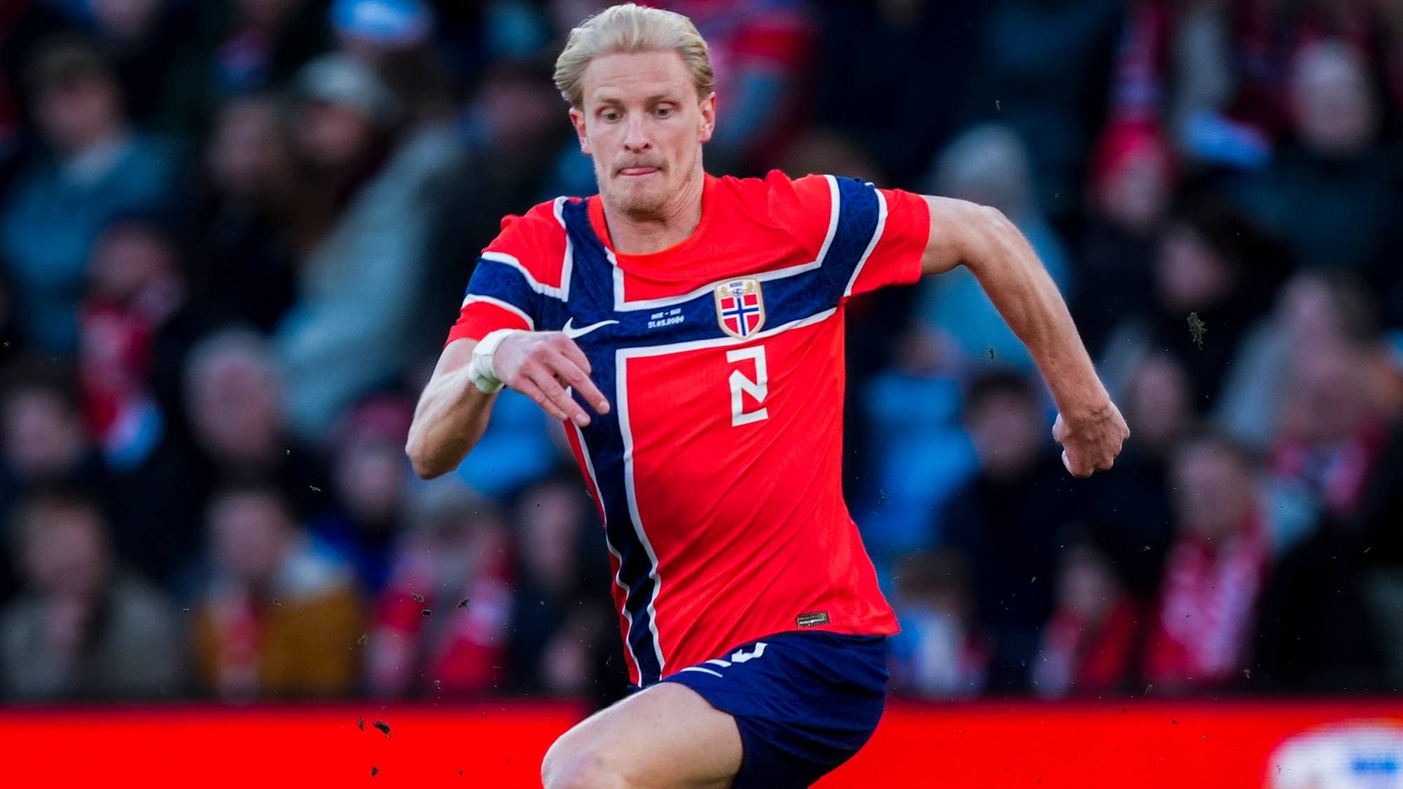 En mann i rød drakt med norsk flagg på brystet spiller fotball. Han er i aksjon på en gressbane med tilskuere i bakgrunnen. Bakgrunnen er en rød vegg med et stort, utydelig emblem på. (Bildebeskrivelsen er laget av en KI-tjeneste)