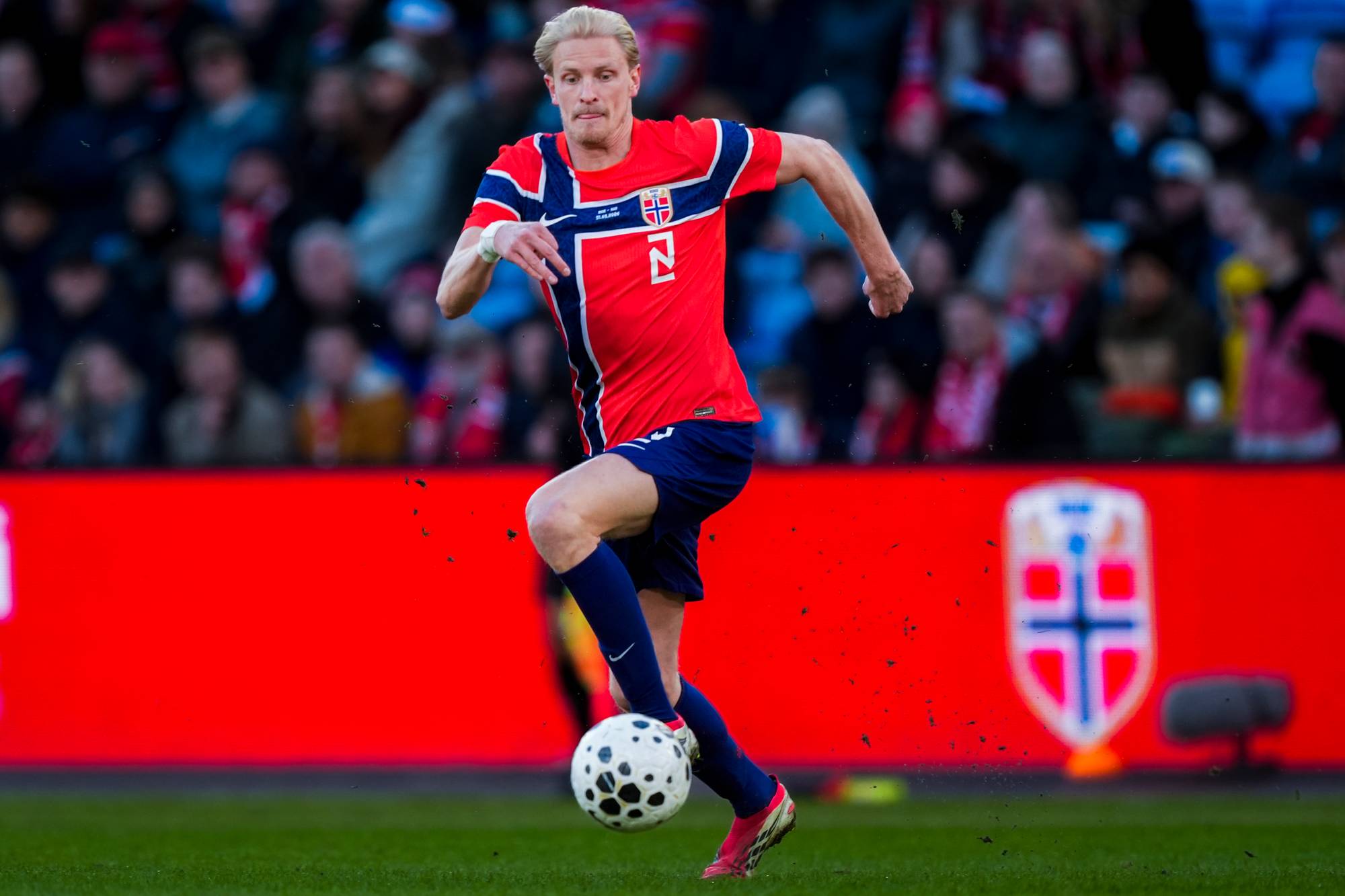 En mann i rød drakt med norsk flagg på brystet spiller fotball. Han er i aksjon på en gressbane med tilskuere i bakgrunnen. Bakgrunnen er en rød vegg med et stort, utydelig emblem på. (Bildebeskrivelsen er laget av en KI-tjeneste)