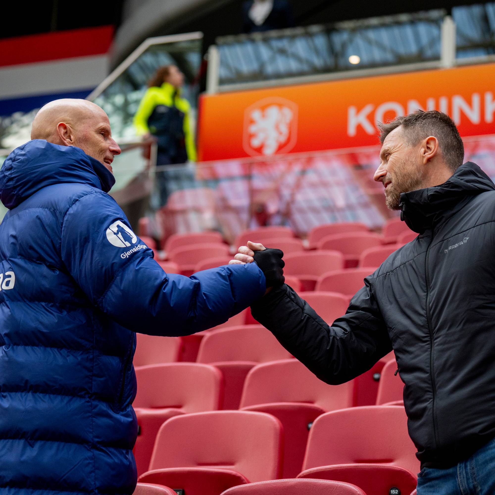 To menn gir hverandre hånden på en tribun. De står på en tribune med røde seter. Bak dem er det et banner med teksten "Koninklijke". (Bildebeskrivelsen er laget av en KI-tjeneste)