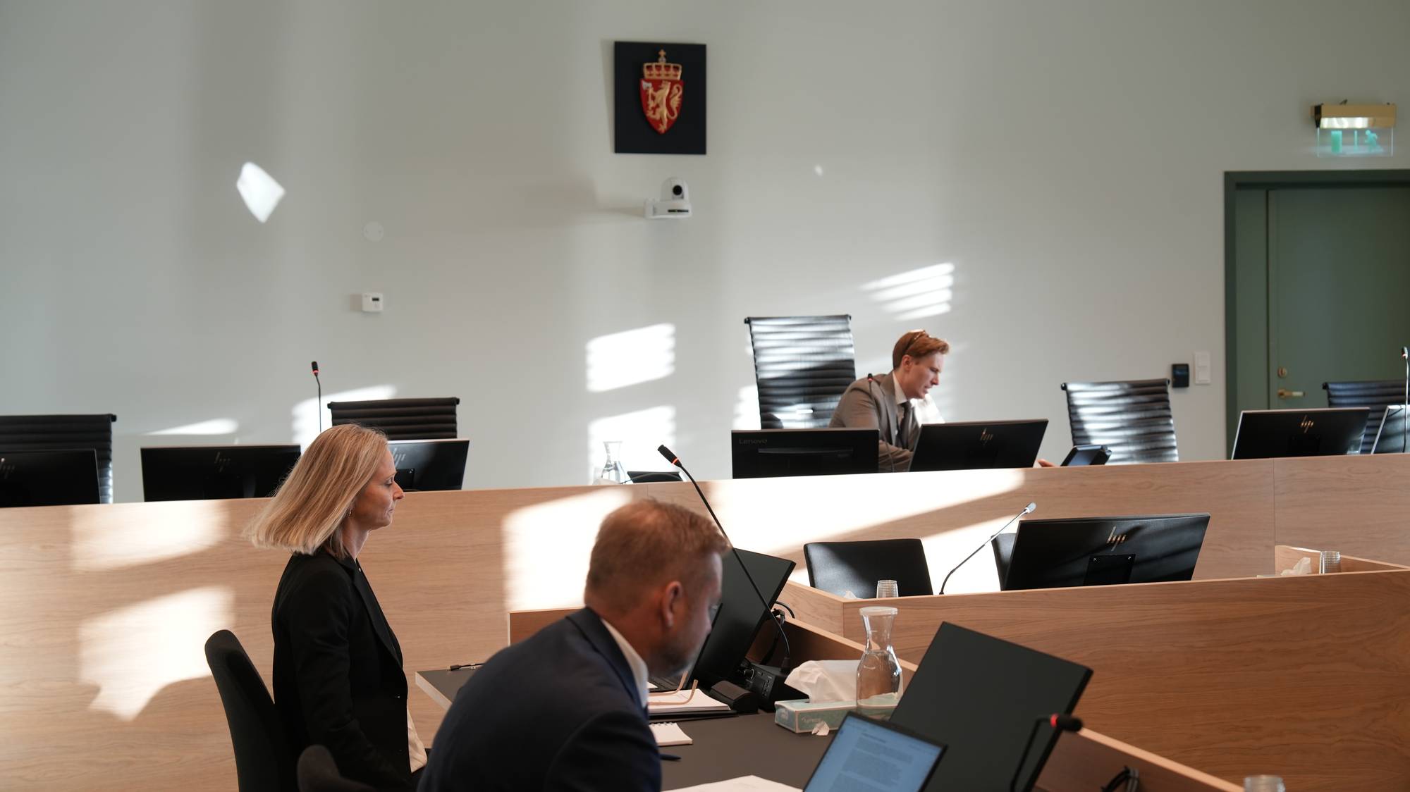 Personer sitter i en rettssal med riksvåpenet på veggen. (Bildebeskrivelsen er laget av en KI-tjeneste)