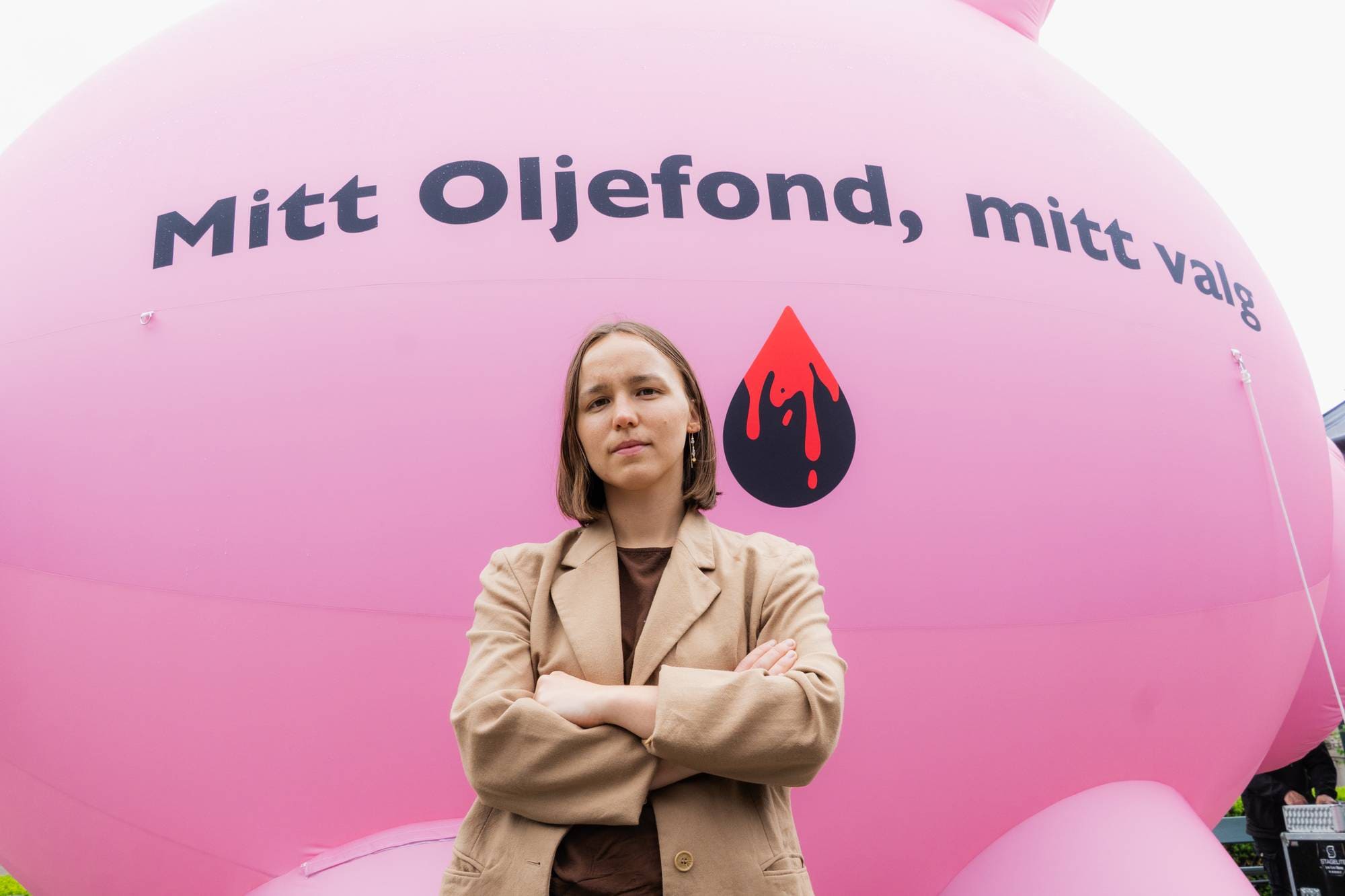 Ung kvinne med alvorlig ansiktsuttrykk og armene i kors står foran en rosa opplåsbar plastgris med teksten "Mitt Oljefond, mitt valg". 