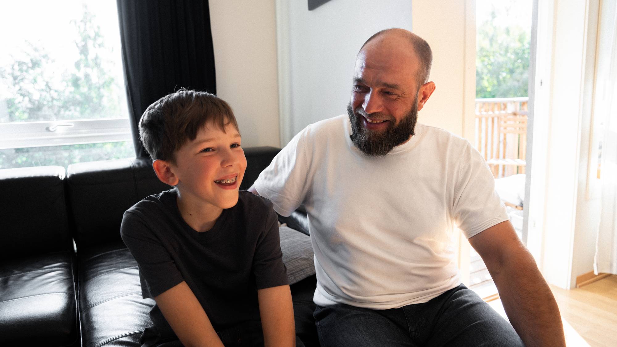 En mann med skjegg og en gutt sitter sammen i en sofa og smiler. 