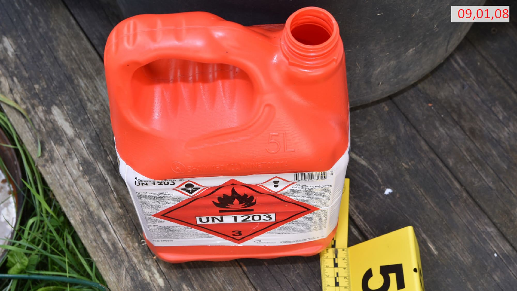 En rød bensinkanne står på et underlag av tre. Man ser litt gress og et plastskilt som er satt opp ved siden av bensinkanna.