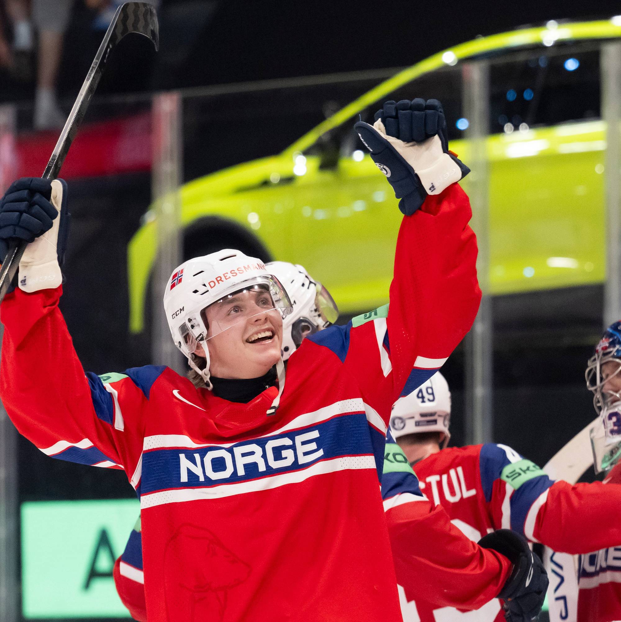 En ishockeyspiller i rød drakt jubler med en stav. Han er omringet av lagkamerater, deriblant en keeper. I bakgrunnen er en gul bil synlig. (Bildebeskrivelsen er laget av en KI-tjeneste)