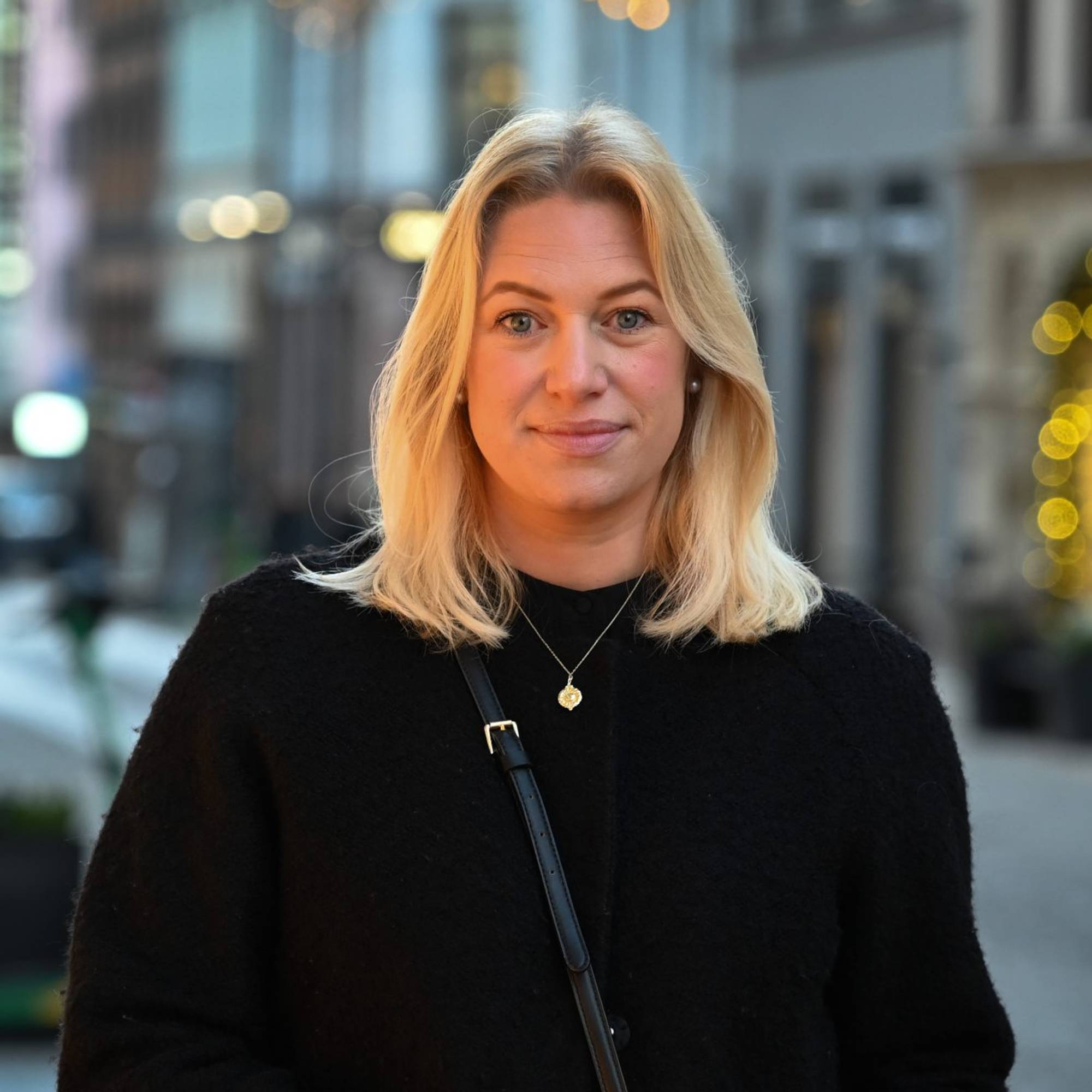 En kvinne ser i kamera. Hun har blondt hår og en svart genser på seg. Hun står i et urbant miljø. 
