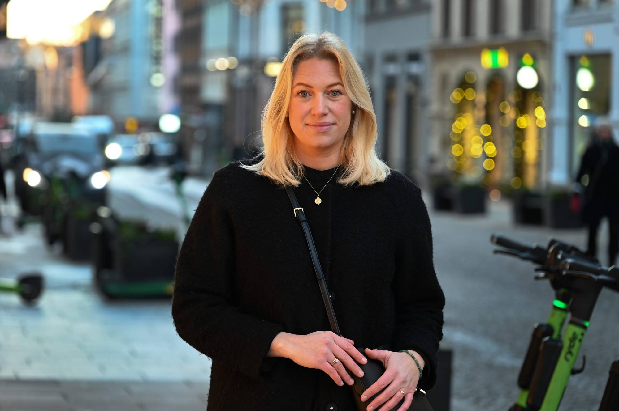 En kvinne ser i kamera. Hun har blondt hår og en svart genser på seg. Hun står i et urbant miljø. 