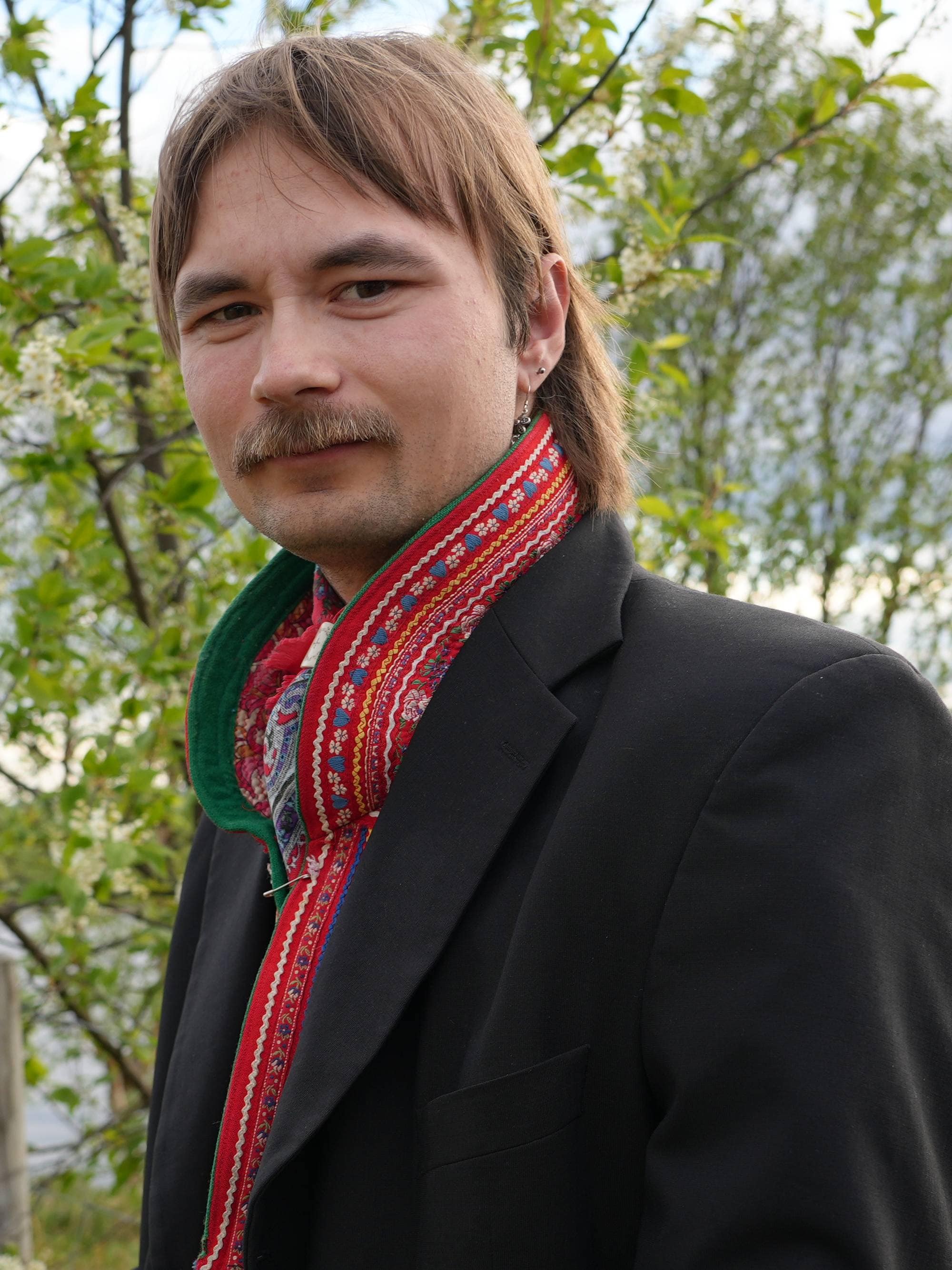 En mann med bart og en fargerik scarf står foran et tre og en innsjø. Han har på seg en mørk dressjakke og en blå skjorte. Bak mannen sees et gammelt gjerde, grønt gress og trær. Været er overskyet med noen solflekker. (Bildebeskrivelsen er laget av en KI-tjeneste)