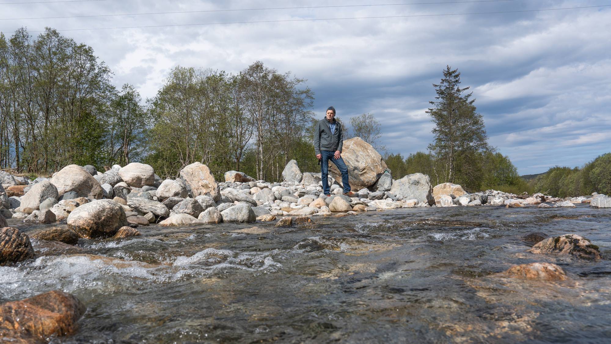 En mann står ved en elv. Mannen er kledd i en mørk jakke og blå jeans. I bakgrunnen er det trær og en grå skydekket himmel. (Bildebeskrivelsen er laget av en KI-tjeneste)