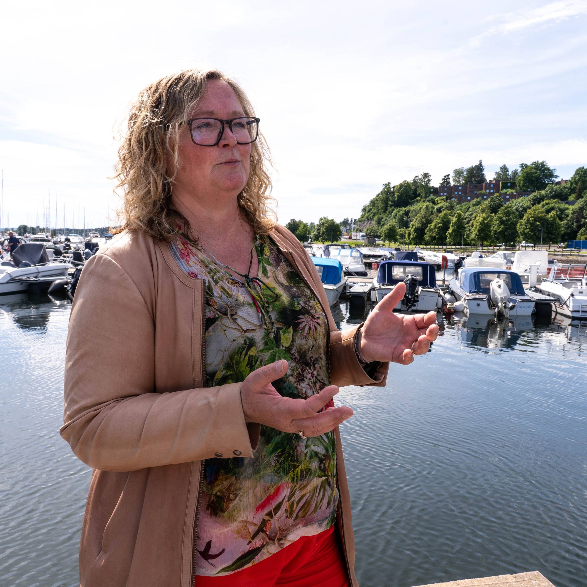 Kvinne med blondt hår og beige skinnjakke med småbåthavn i bakgrunnen