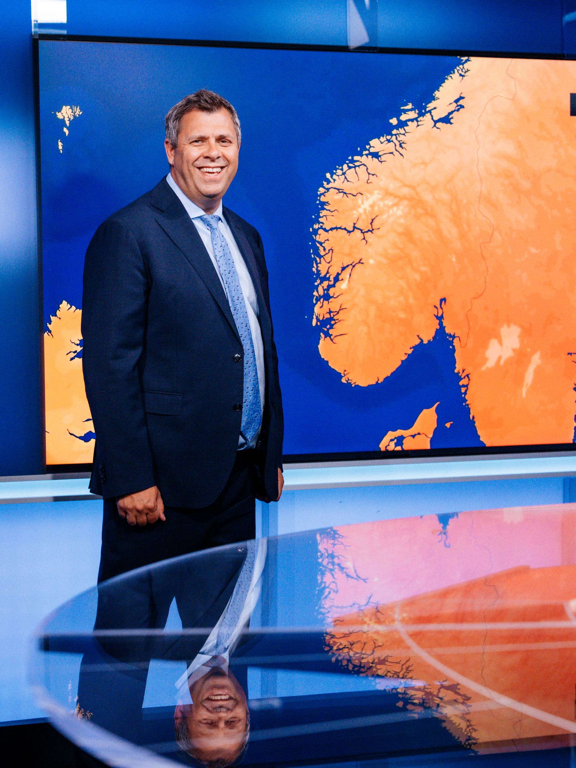 Mann i dress står i et tv-studio foran et kart over Norge. (Bildebeskrivelsen er laget av en KI-tjeneste)