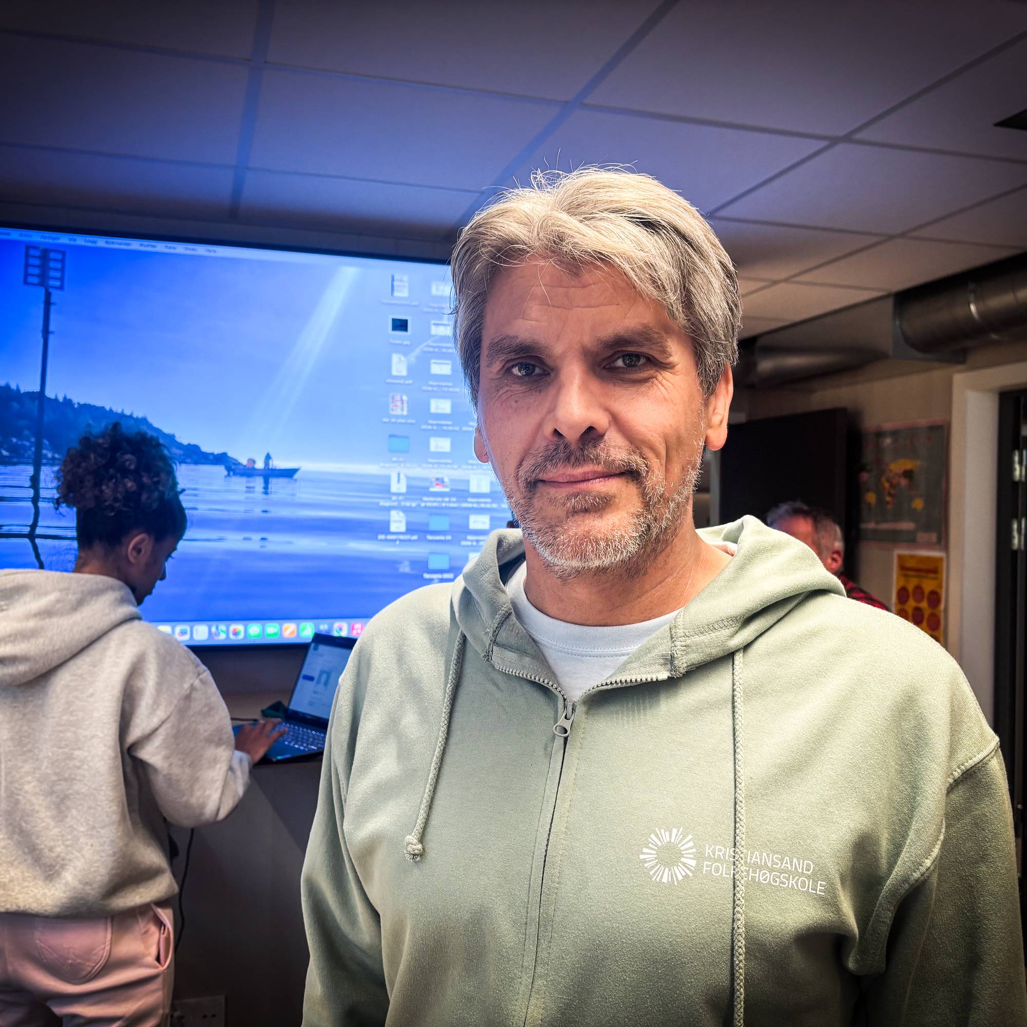 En mann i lys grønn hettegenser står foran en skjerm i et rom med folk i bakgrunnen. (Bildebeskrivelsen er laget av en KI-tjeneste)