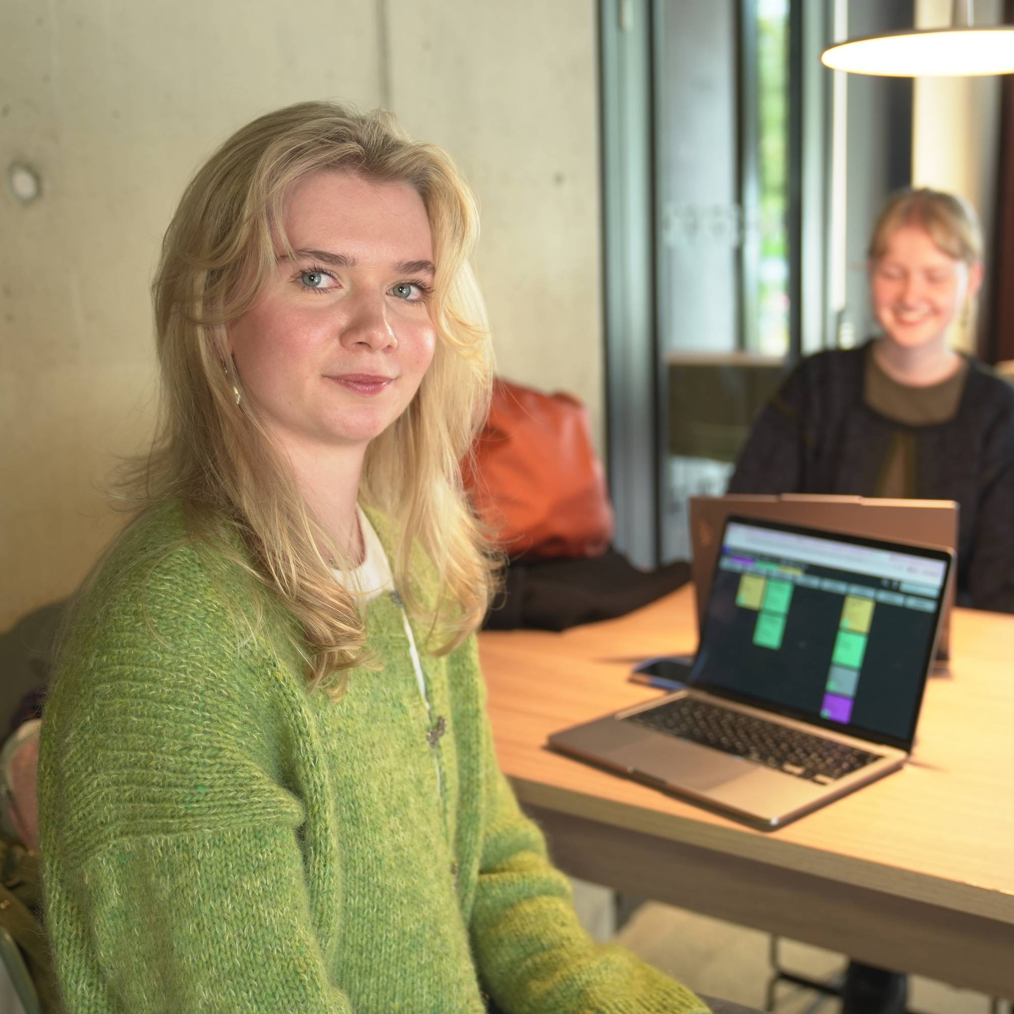 Ung kvinne i grønn genser sitter foran en PC. Hun ser inn i kameraet.
