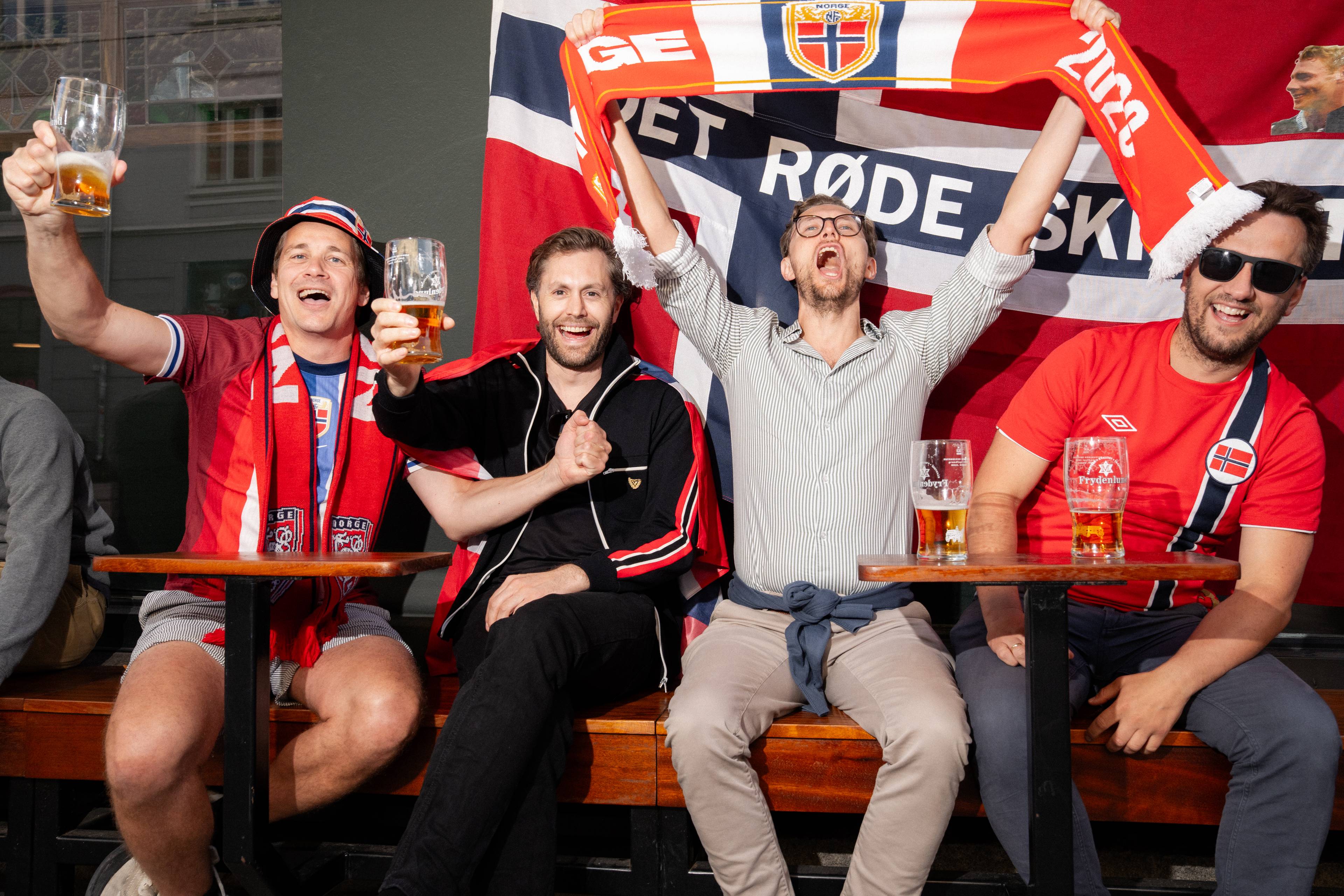 Fire menn sitter bak et bord med ølglass, iført norske supporterklær. En stor norsk fane er bak dem. Mennene holder ølglass i været og ler. (Bildebeskrivelsen er laget av en KI-tjeneste)