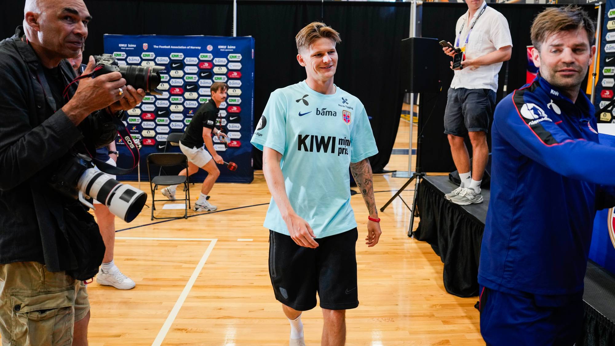 En mann i lys blå t-skjorte går mot kamera. Han har svart shorts og hvite sokker. En fotograf til venstre holder et stort kamera. En annen mann i blå genser står til høyre for t-skjortemannen. Bak dem er et bakteppe med logoer. (Bildebeskrivelsen er laget av en KI-tjeneste)