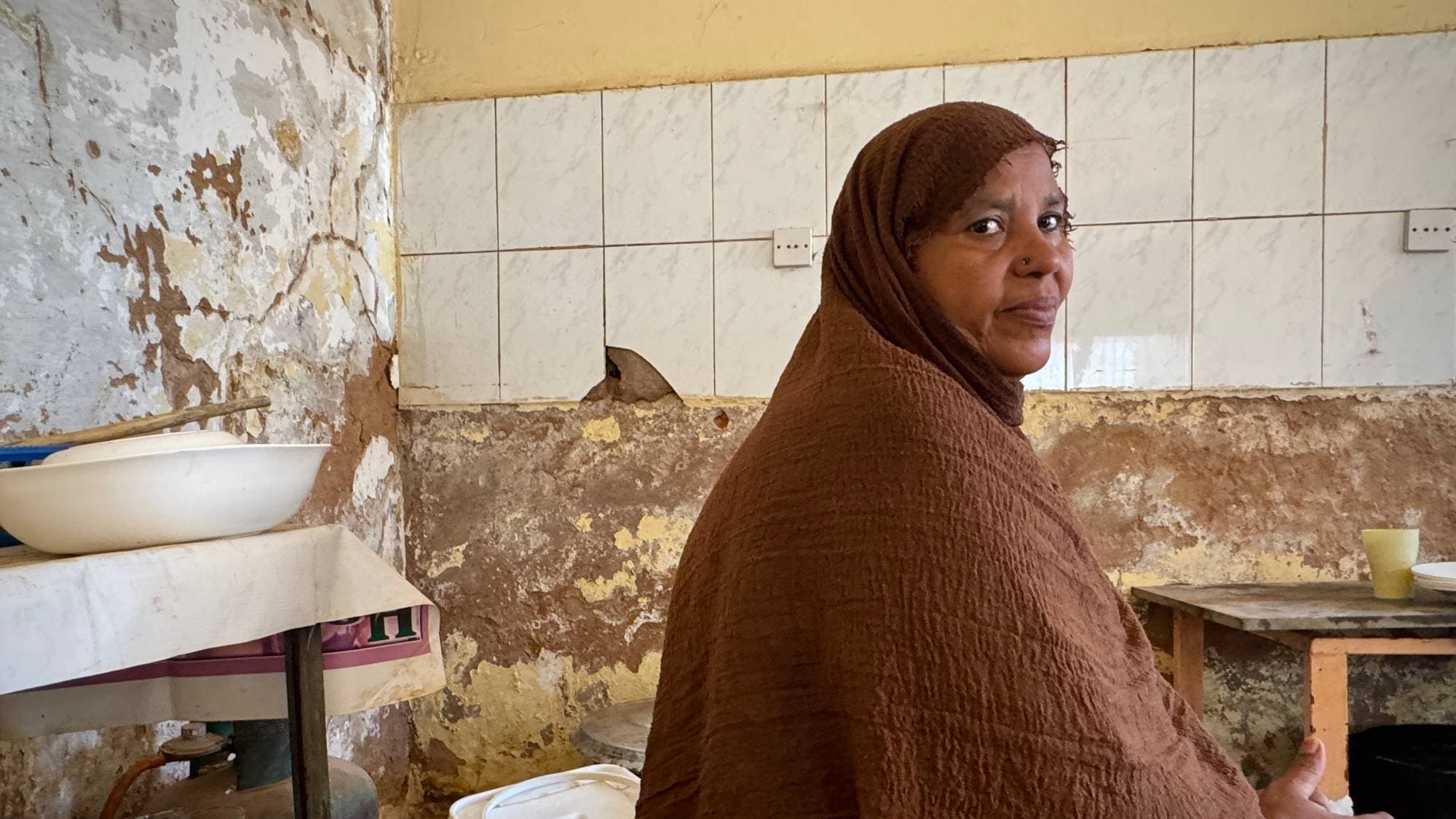 Ei sudansk kvinne sit på eit kjøkken og kikkar mot kamera. Ho har på seg ein brun hijab og har eit lite smil.