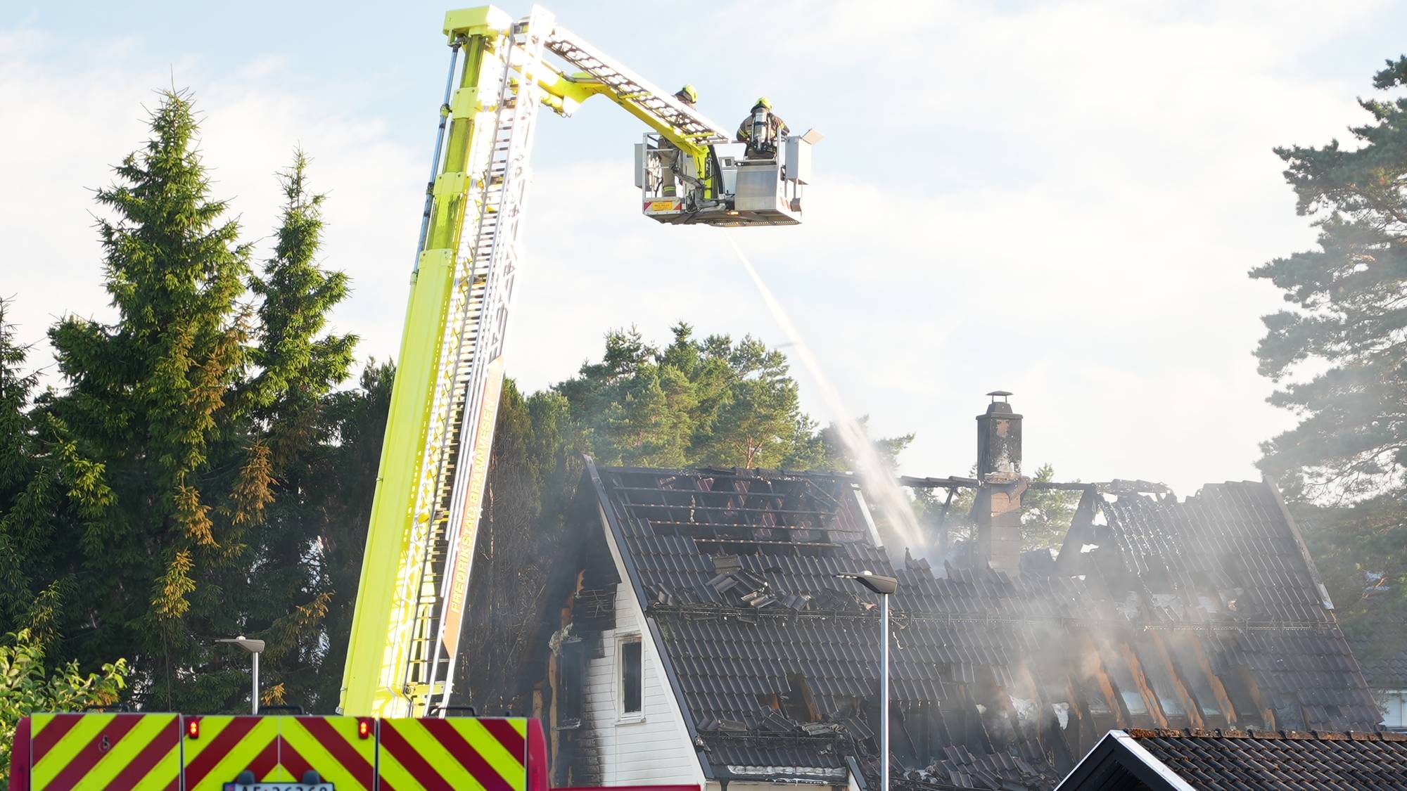 Brannvesen slukker brann i en enebolig med liftbil. En brannkonstabel går mot huset. Politibil står nær. (Bildebeskrivelsen er laget av en KI-tjeneste)