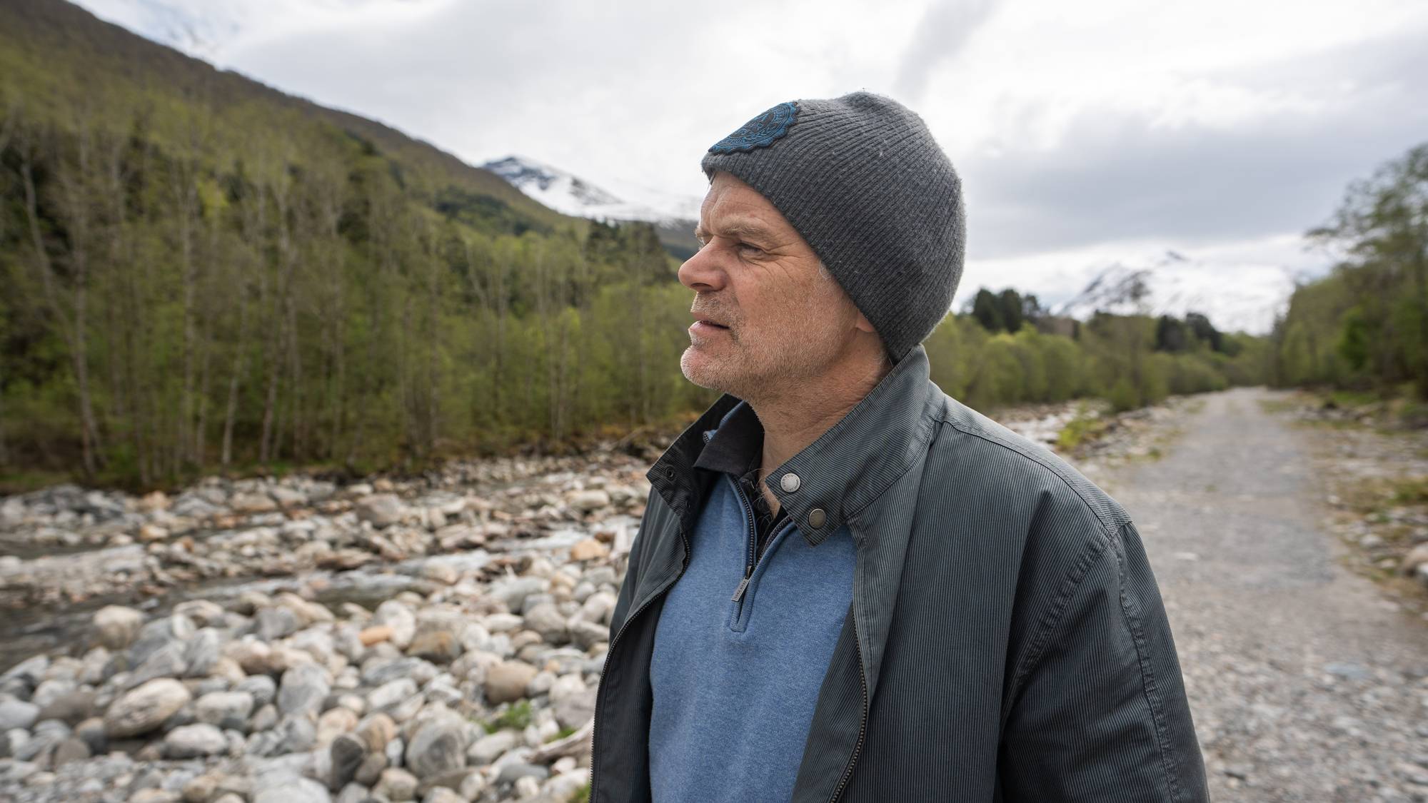 En mann i hatt og jakke ser mot venstre. Bak ham er en elv og et fjellandskap. En grusvei går inn i bildet fra høyre. (Bildebeskrivelsen er laget av en KI-tjeneste)