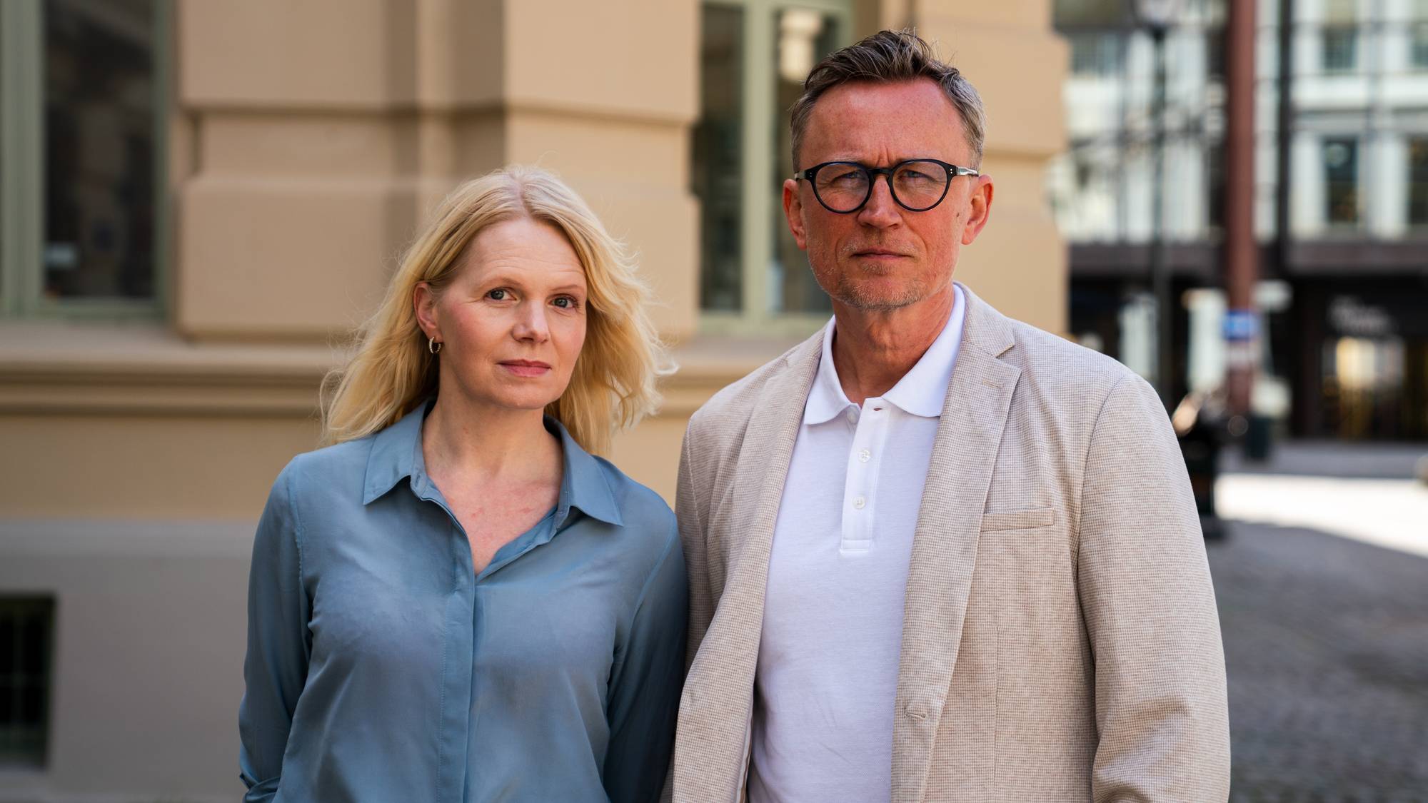 En kvinne i blå skjorte står ved siden av en mann i hvit pirquet-skjorte med beige blazer over. Kvinnen har langt, blondt hår som fragler i vinden og mannen har kort mørkt hår og briller. Begge ser inn i kamera med en nøytral mine. 