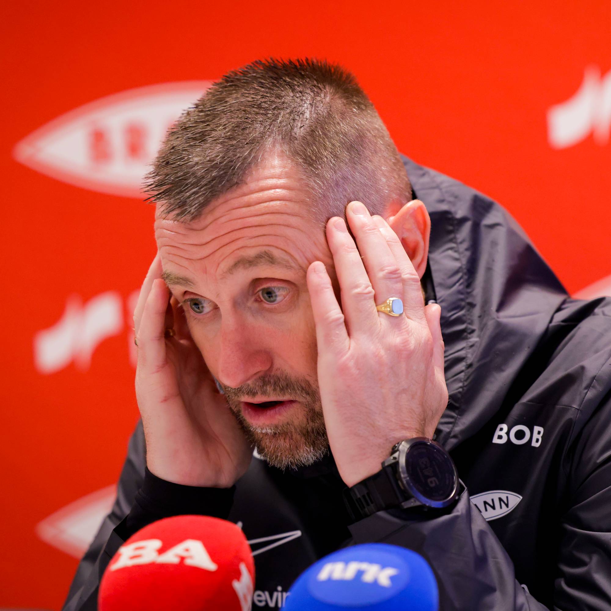 Trener Eirik Horneland holder seg til hodet under en pressekonferanse med Brann-logoer i bakgrunnen. (Bildebeskrivelsen er laget av en KI-tjeneste)