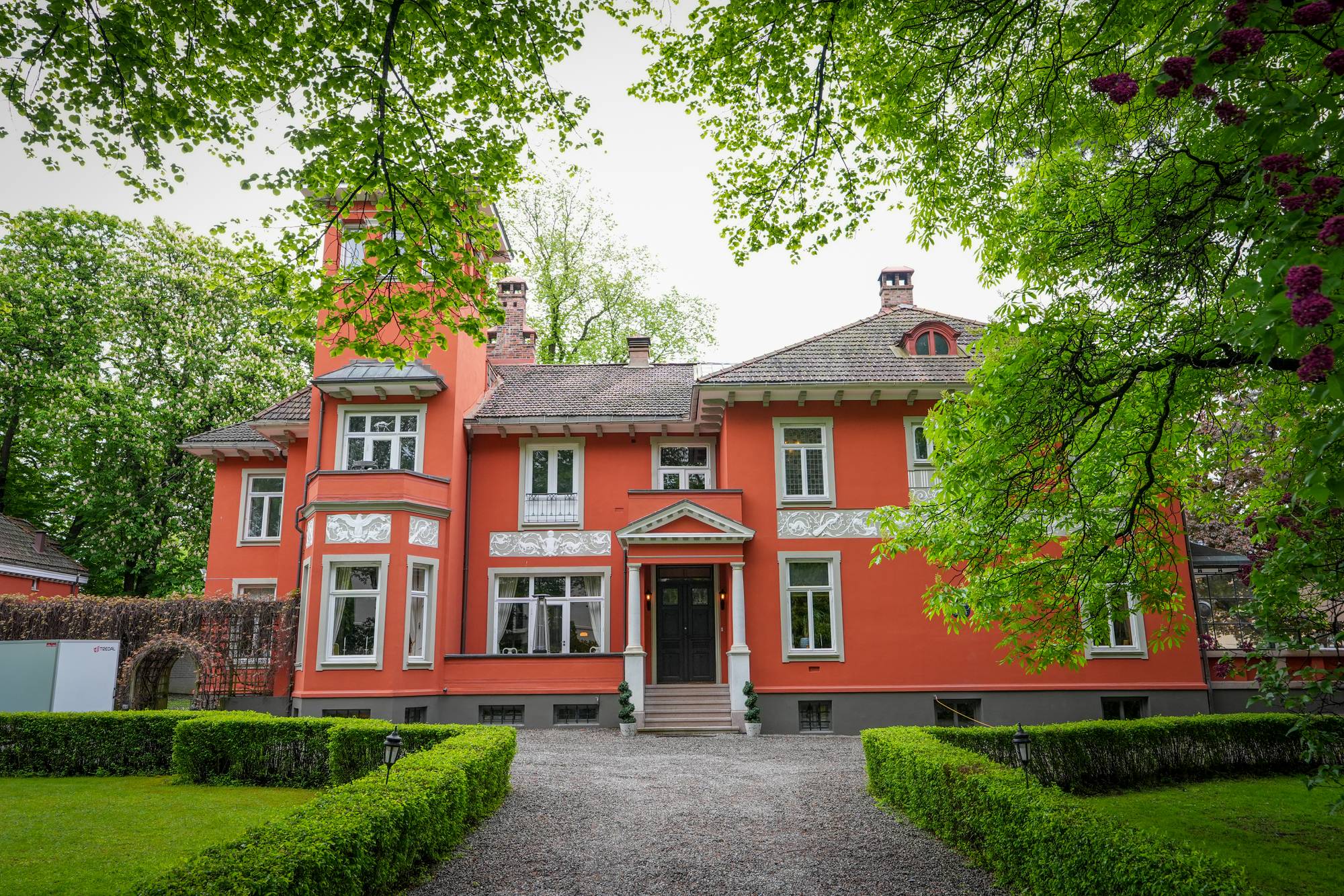 Stor rød villa med tårn og hage foran. Gruslagt adkomstvei omgitt av lave hekker. (Bildebeskrivelsen er laget av en KI-tjeneste)
