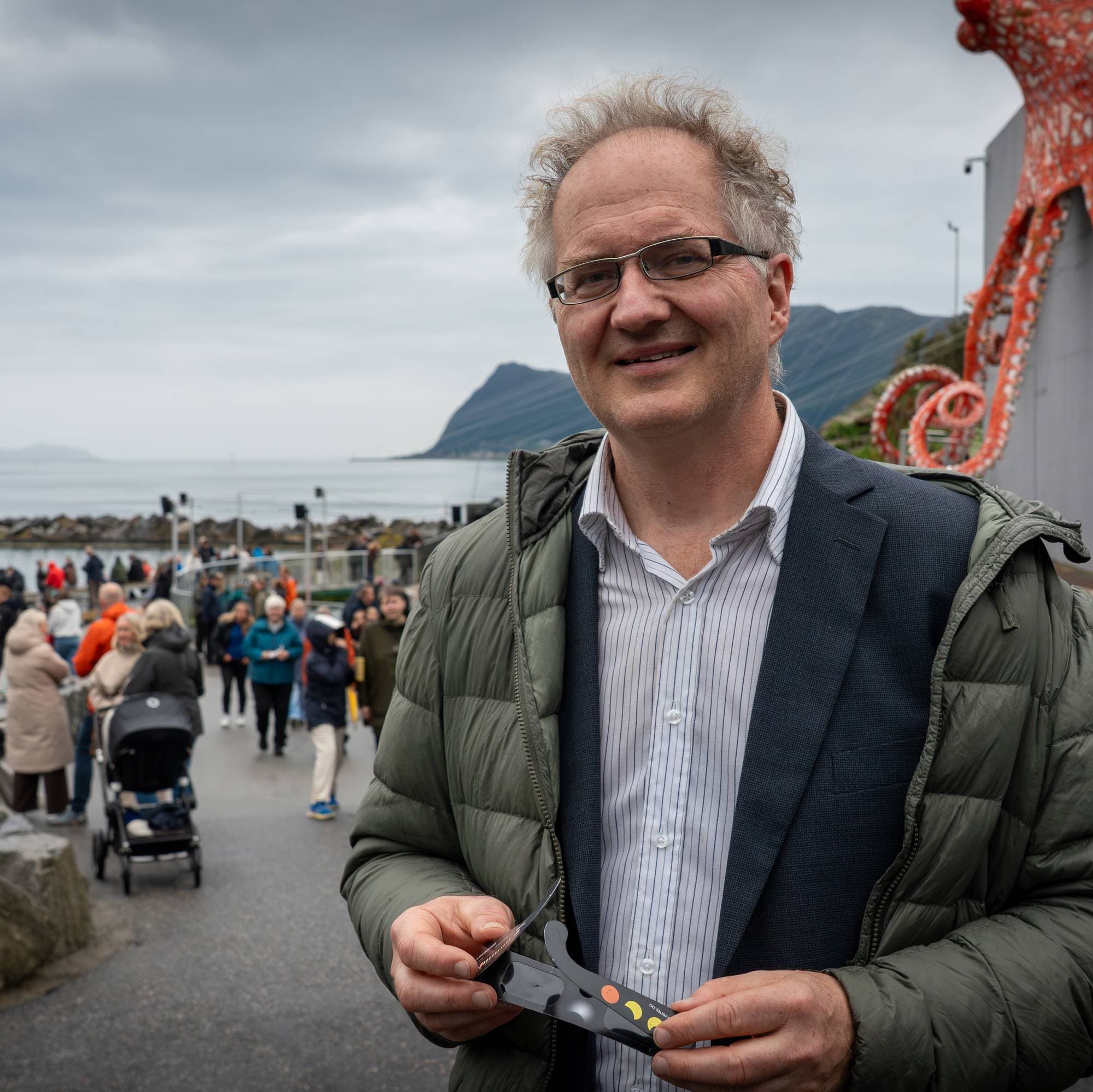 En mann holder solbriller foran en blekksprutskulptur ved sjøen mens andre folk går i bakgrunnen. (Bildebeskrivelsen er laget av en KI-tjeneste)