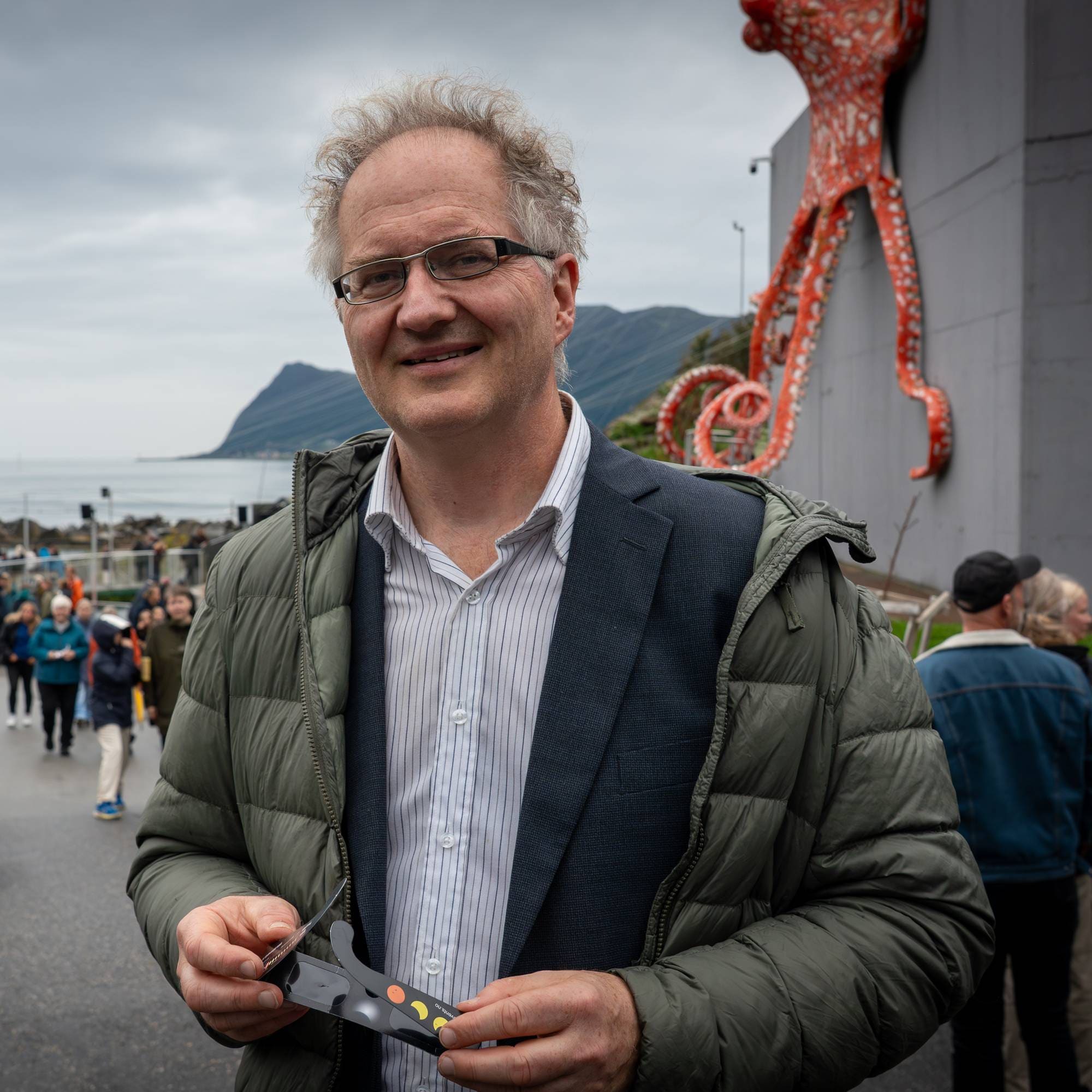 En mann holder solbriller foran en blekksprutskulptur ved sjøen mens andre folk går i bakgrunnen. (Bildebeskrivelsen er laget av en KI-tjeneste)