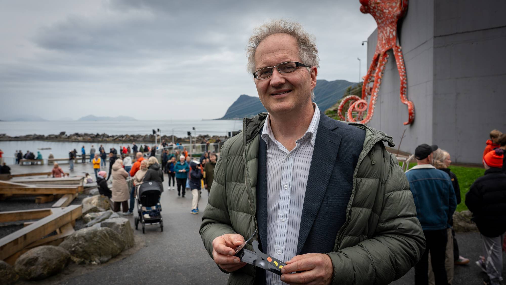 En mann holder solbriller foran en blekksprutskulptur ved sjøen mens andre folk går i bakgrunnen. (Bildebeskrivelsen er laget av en KI-tjeneste)