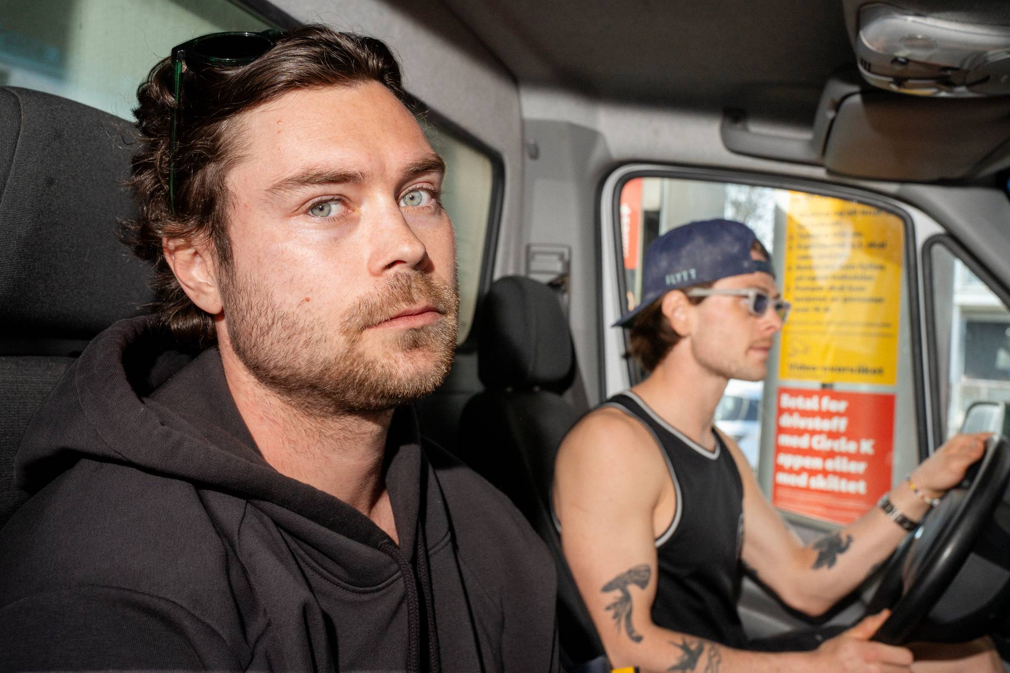 To menn sitter i en varebil. Den ene mannen sitter i passasjersetet og ser mot kamera. Den andre mannen sitter i førersetet og ser ut av vinduet mens han holder på rattet. Utenfor bilen er det en gul og rød plakat med tekst. (Bildebeskrivelsen er laget av en KI-tjeneste)