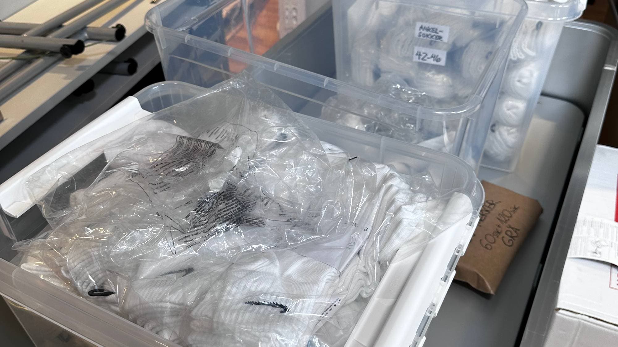 Klarplastbokser med hvite klær står på et metallunderlag. Boksene har etiketter med tekst og tall. I bakgrunnen sees pappesker og et rødt trillebord. Omgivelsene er et lager eller et oppbevaringsrom. (Bildebeskrivelsen er laget av en KI-tjeneste)
