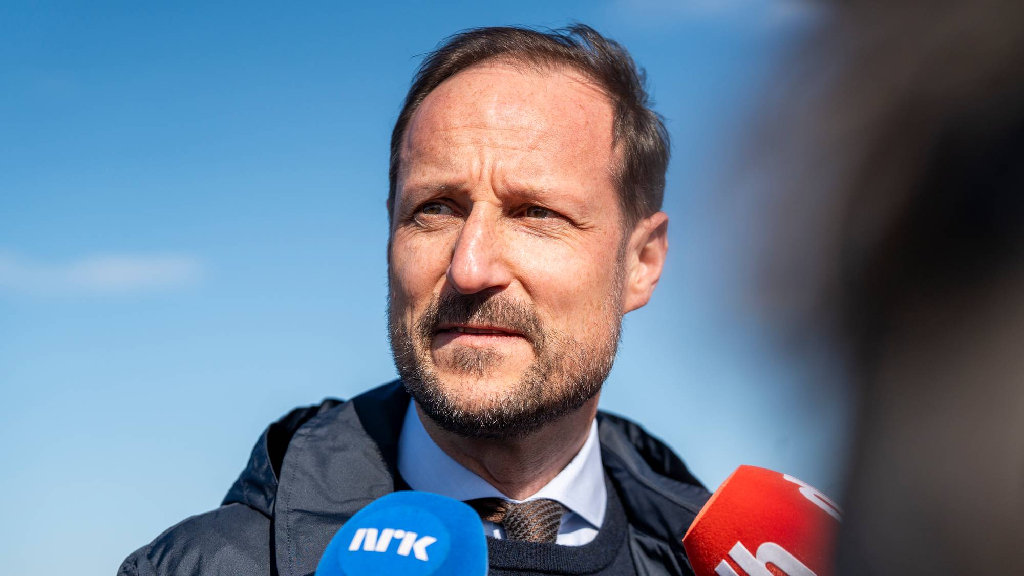 Kronprins Haakon blir intervjuet av journalister med mikrofoner utendørs i solskinn.