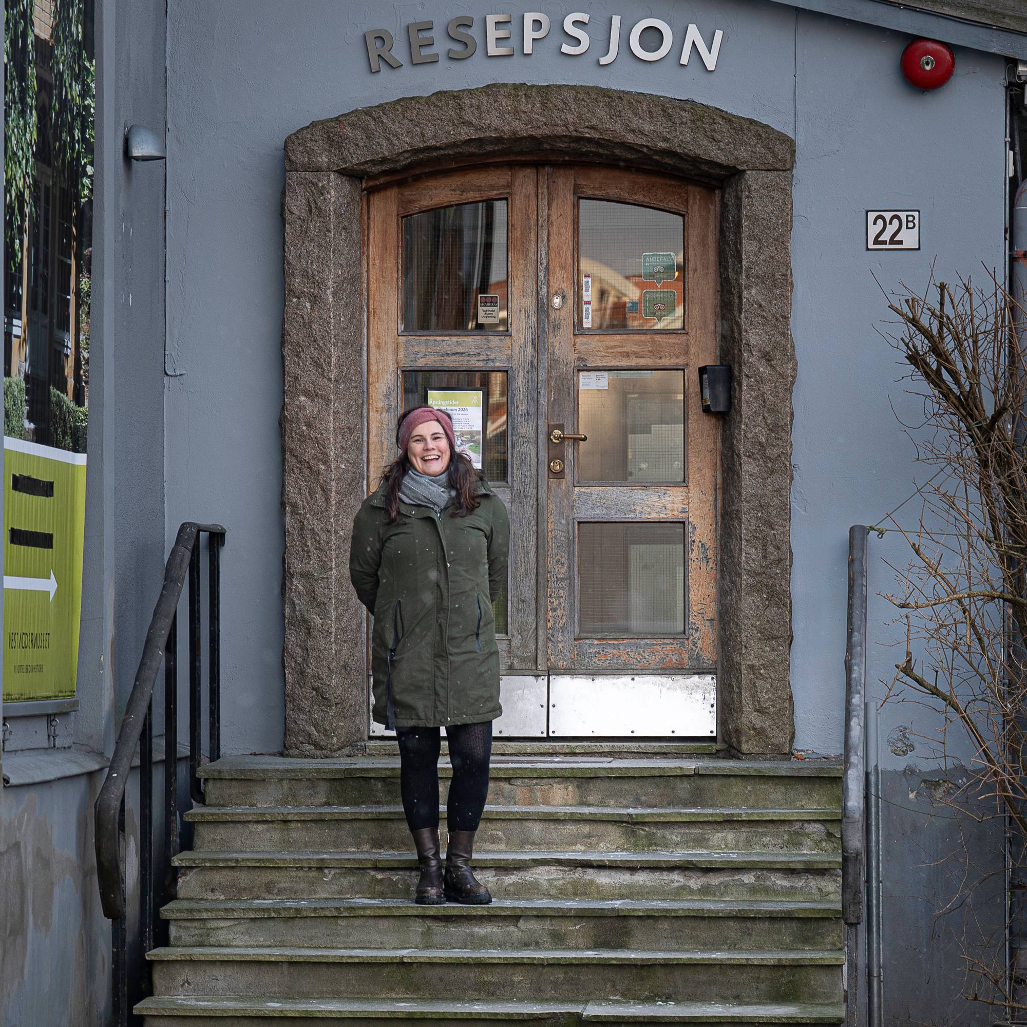 En kvinne smiler i en trapp foran en gammel dør. Over døra står det Resepsjon. 