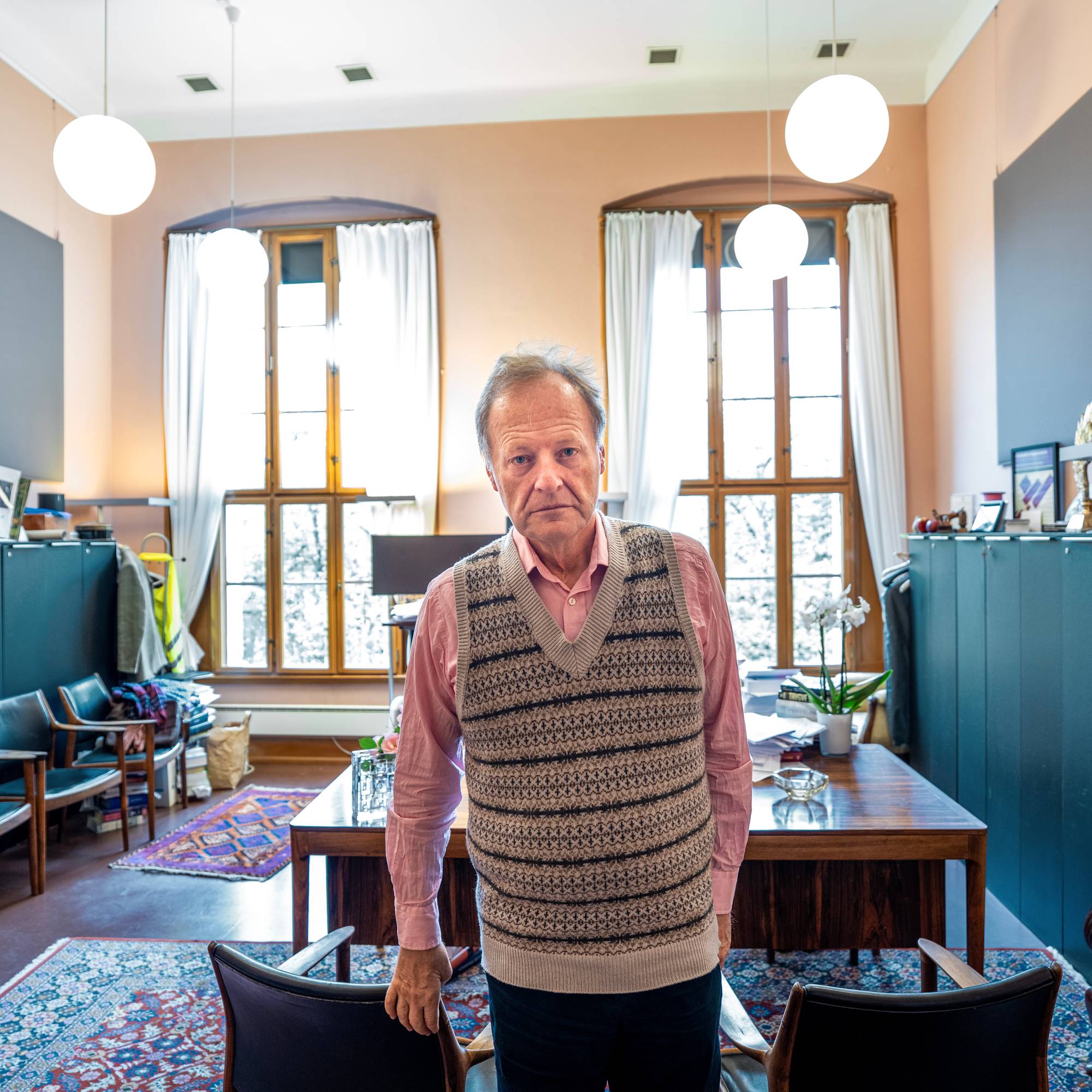En eldre mann med strikkevest står foran et skrivebord på et kontor med store vinduer. (Bildebeskrivelsen er laget av en KI-tjeneste)