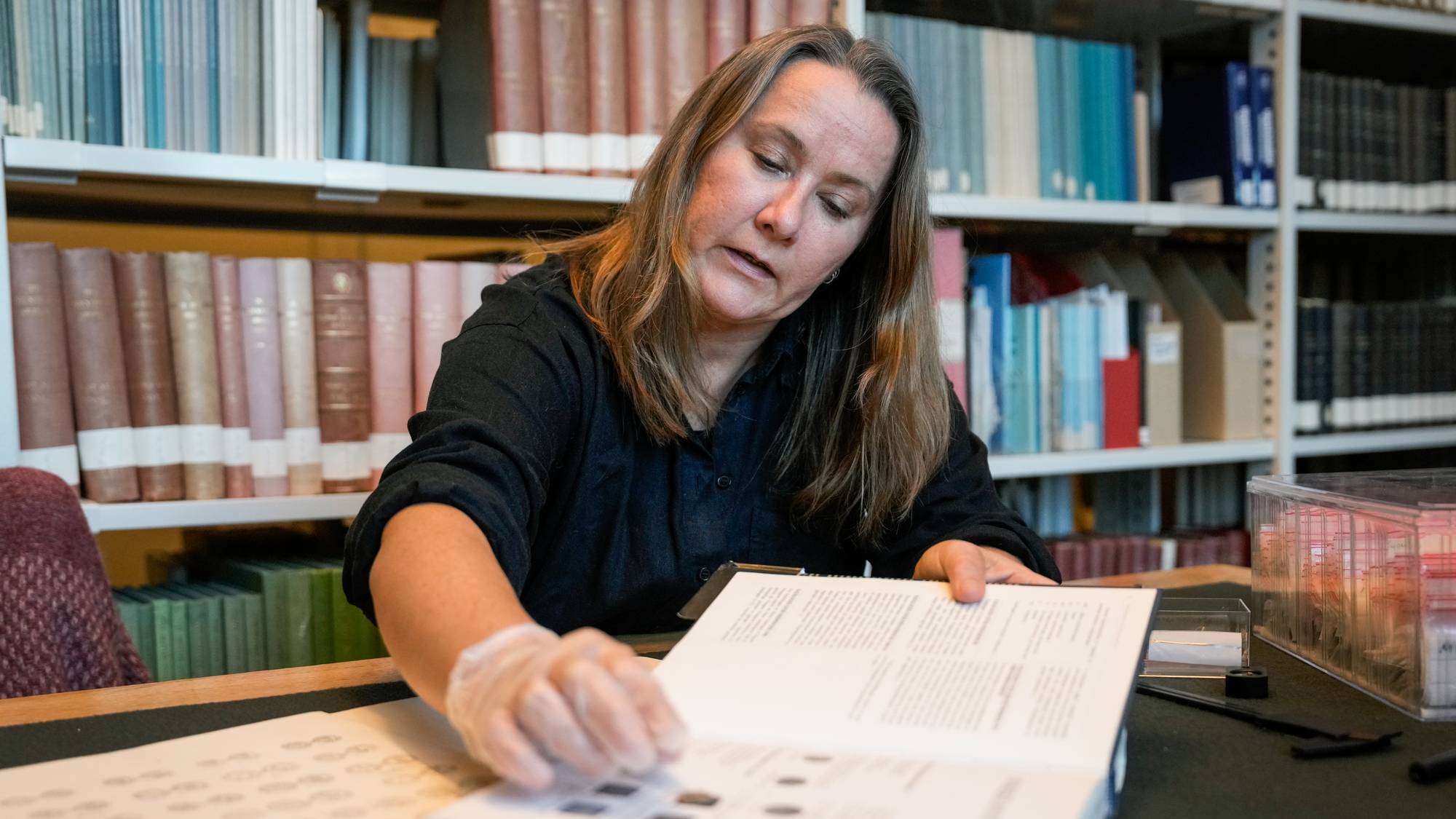 Kvinne med plasthanske blar i en bok på et bibliotek eller arkiv med bokhyller i bakgrunnen. 