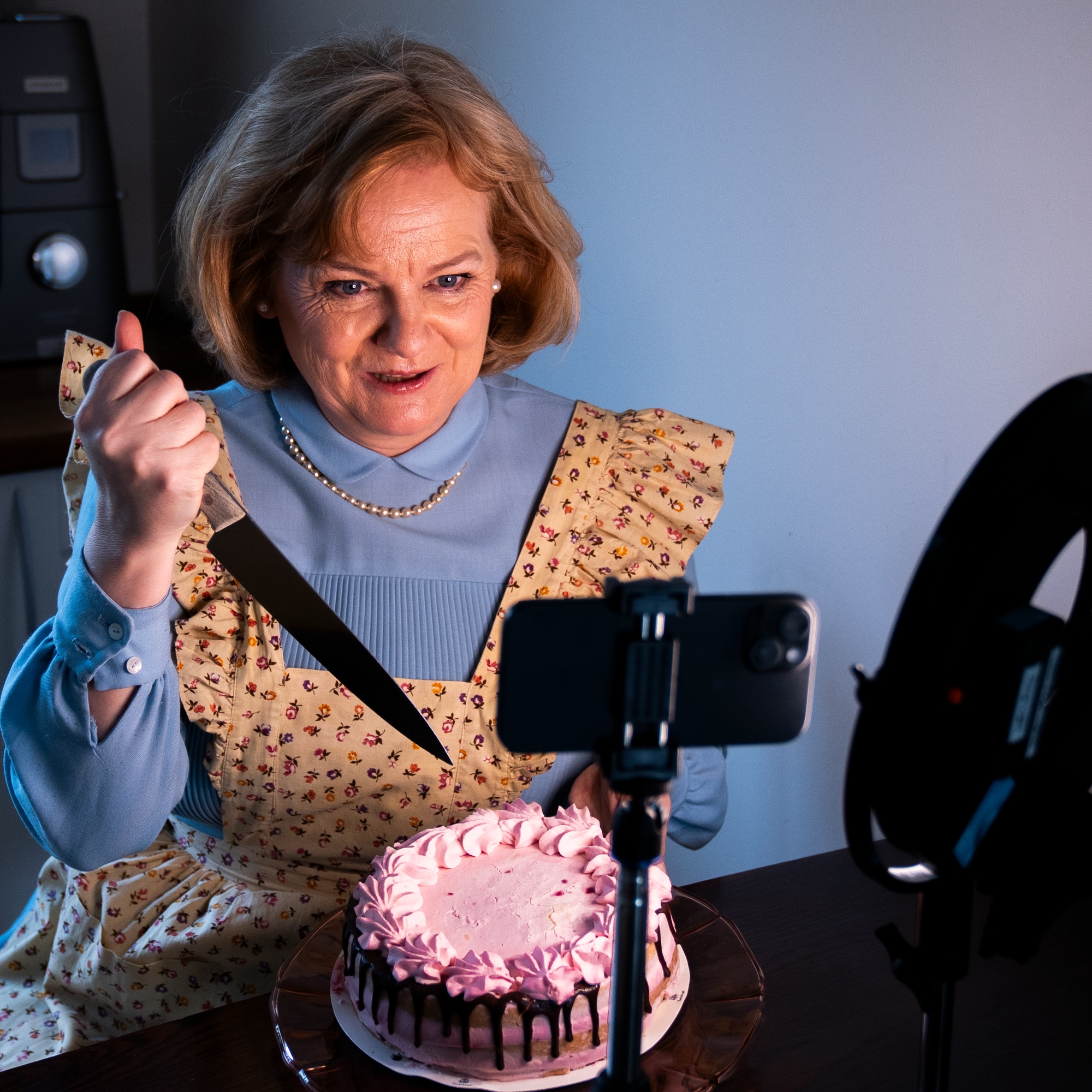 En kvinne i husmorkostyme med et morderisk blikk holder en blodig, dryppende kniv over en pyntet kake i et kjøkken