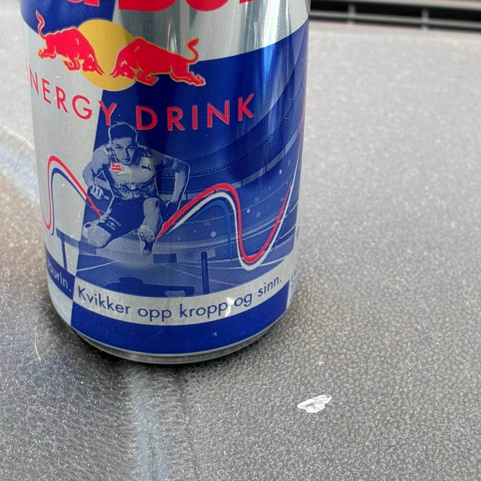 En boks med Red Bull energidrikk plassert på en grå flate. Illustrasjon av Karsten Warholm på siden. (Bildebeskrivelsen er laget av en KI-tjeneste)