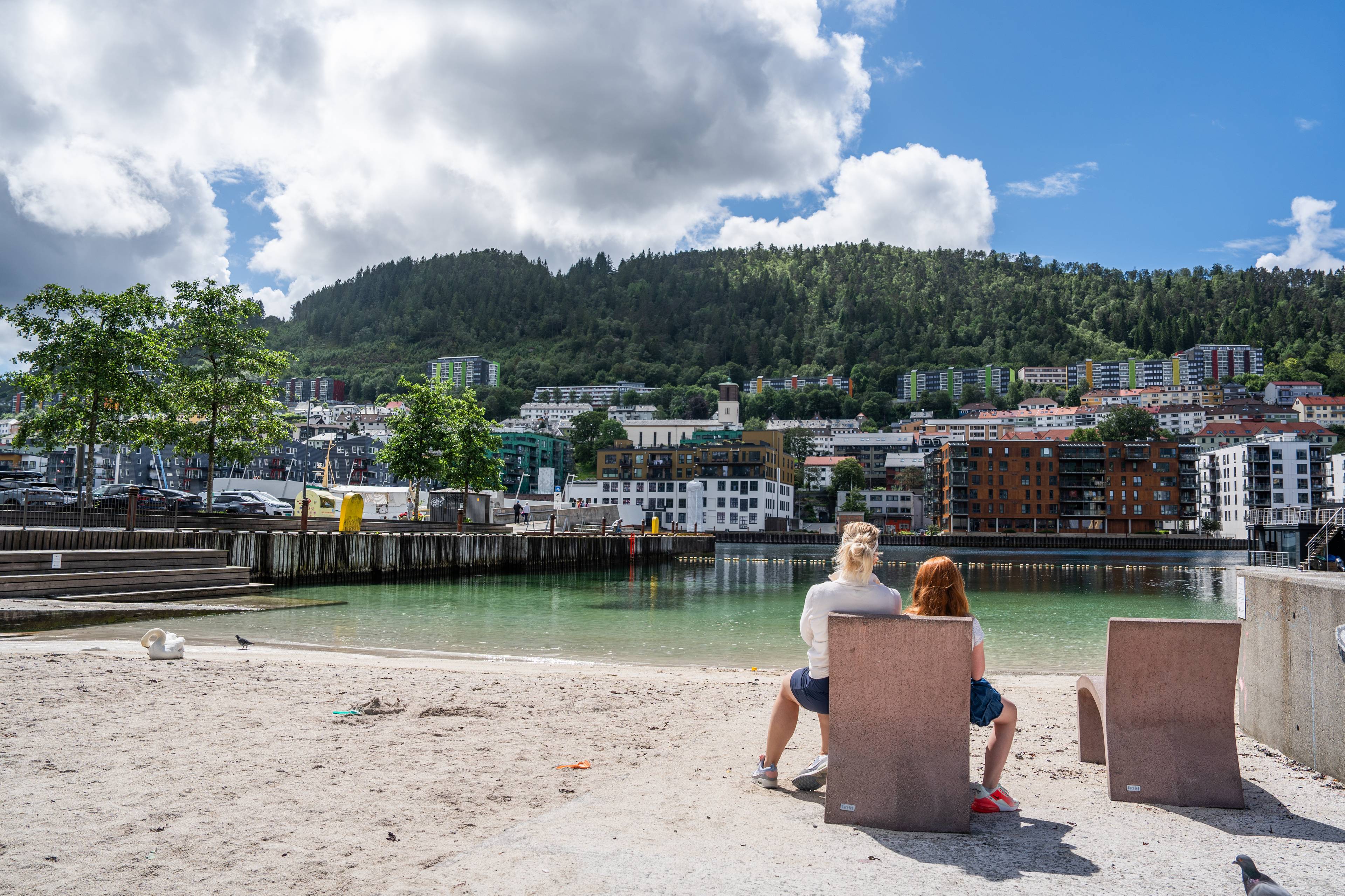 To personer sitter på stoler ved en strand med utsikt mot bebyggelse og skogkledd ås i Bergen. (Bildebeskrivelsen er laget av en KI-tjeneste)