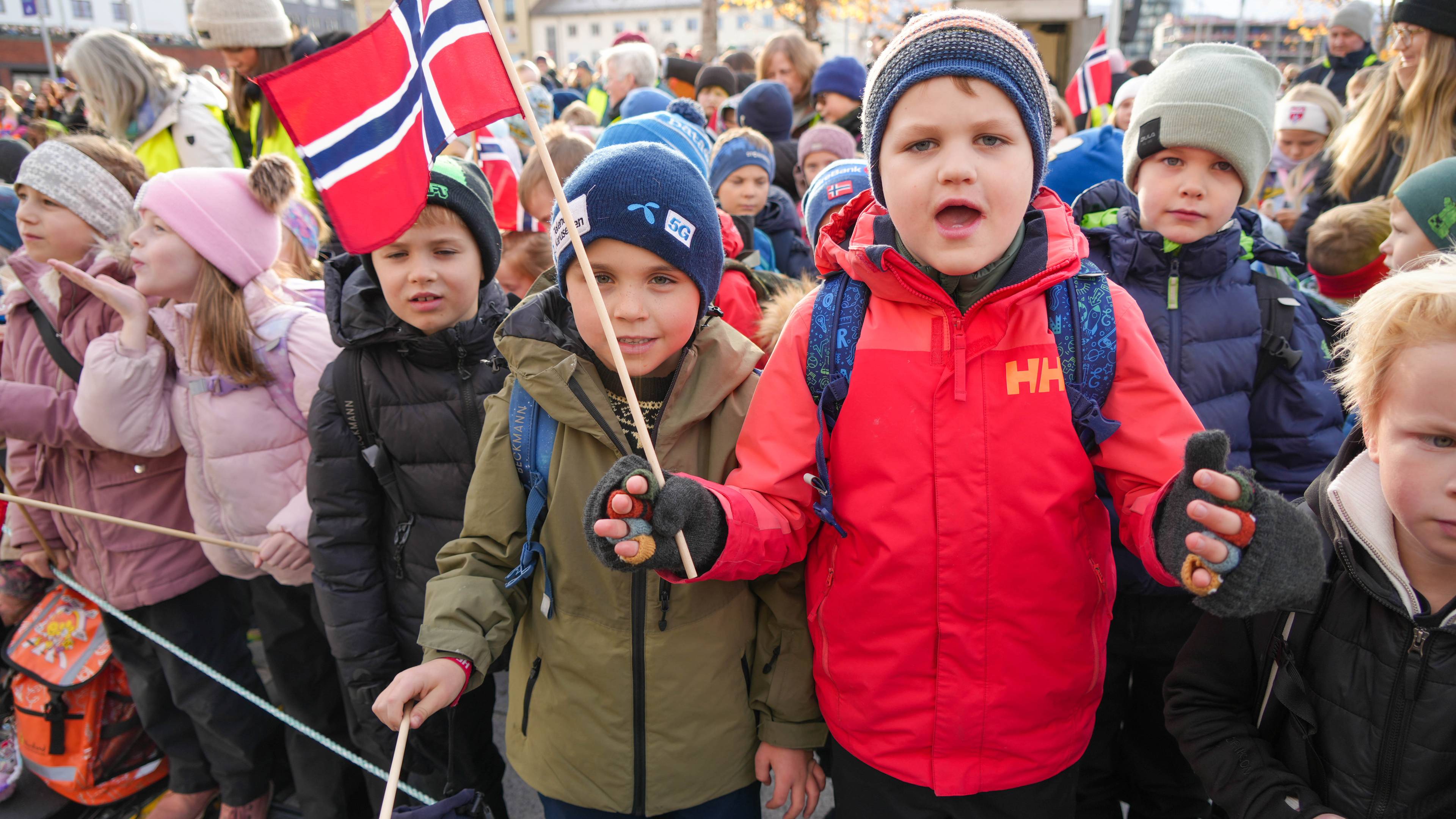 Flere skolebarn med boblejakker og lue på veiver med flagg og venter på kongen.