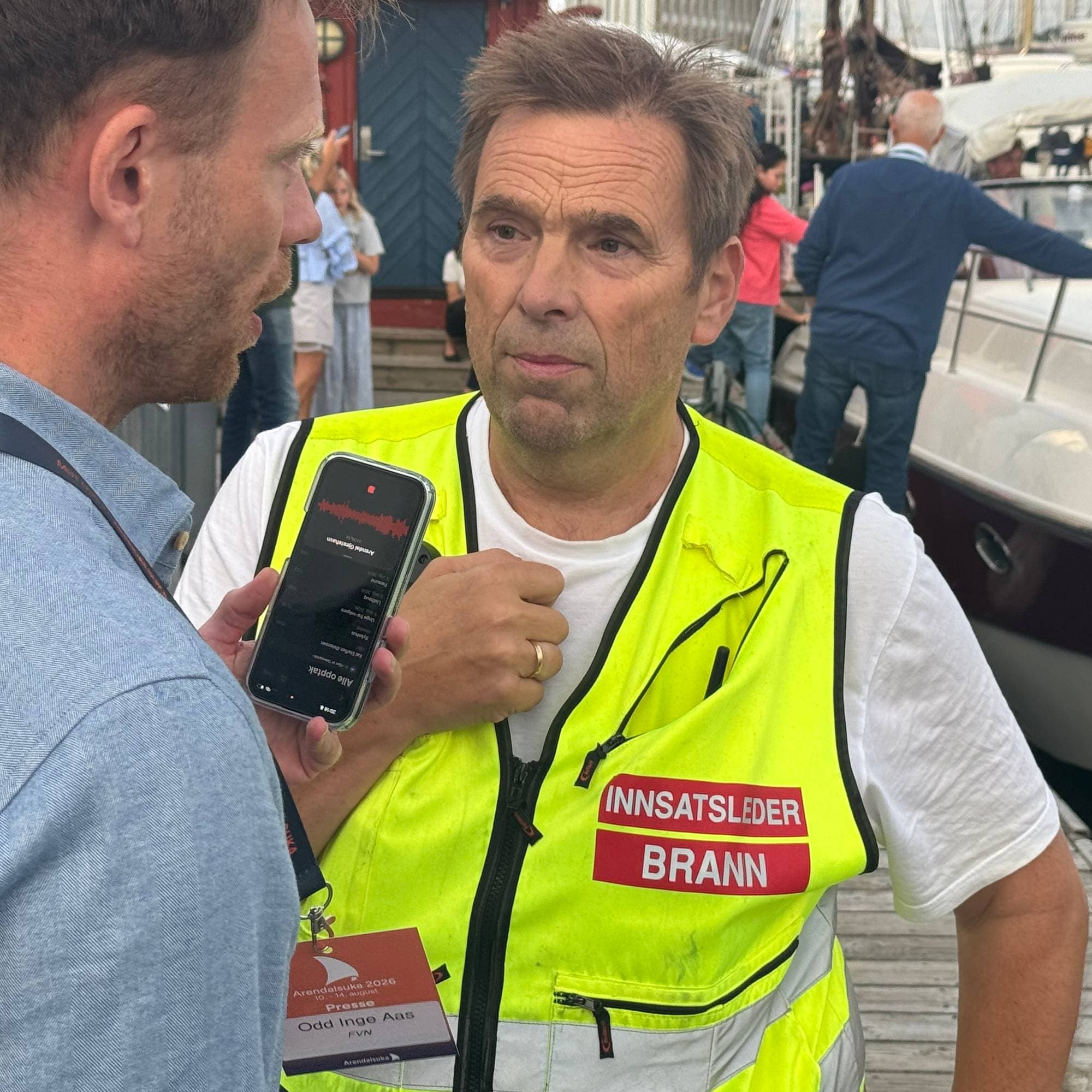 Journalist intervjuer innsatsleder fra brannvesenet i gul refleksvest ved en båthavn. (Bildebeskrivelsen er laget av en KI-tjeneste)