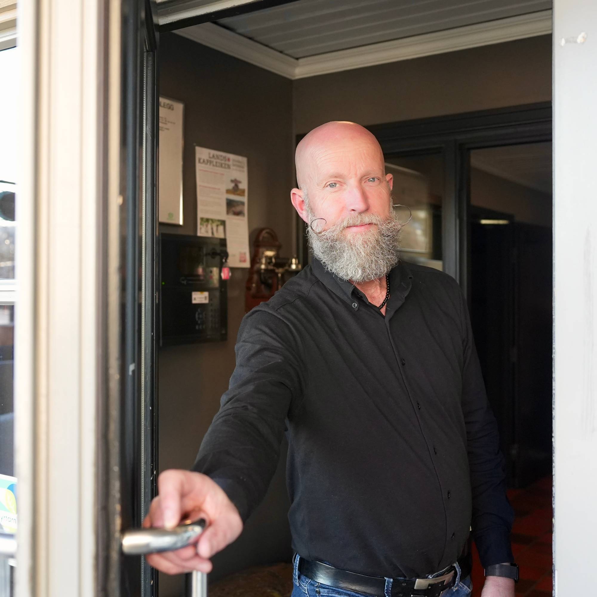 En mann med svart skjorte, skallet og med gråt skjegg holder en dør åpen. 