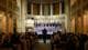 NTB En stor gruppe mennesker synger på en scene i en kirke. Fremst står et barnekor iført hvite skjorter med blå krager, og de er dirigert av en mann. Bak dem er en gruppe voksne menn i mørke dresser. Publikum sitter på benker og ser mot scenen. (Bildebeskrivelsen er laget av en KI-tjeneste)