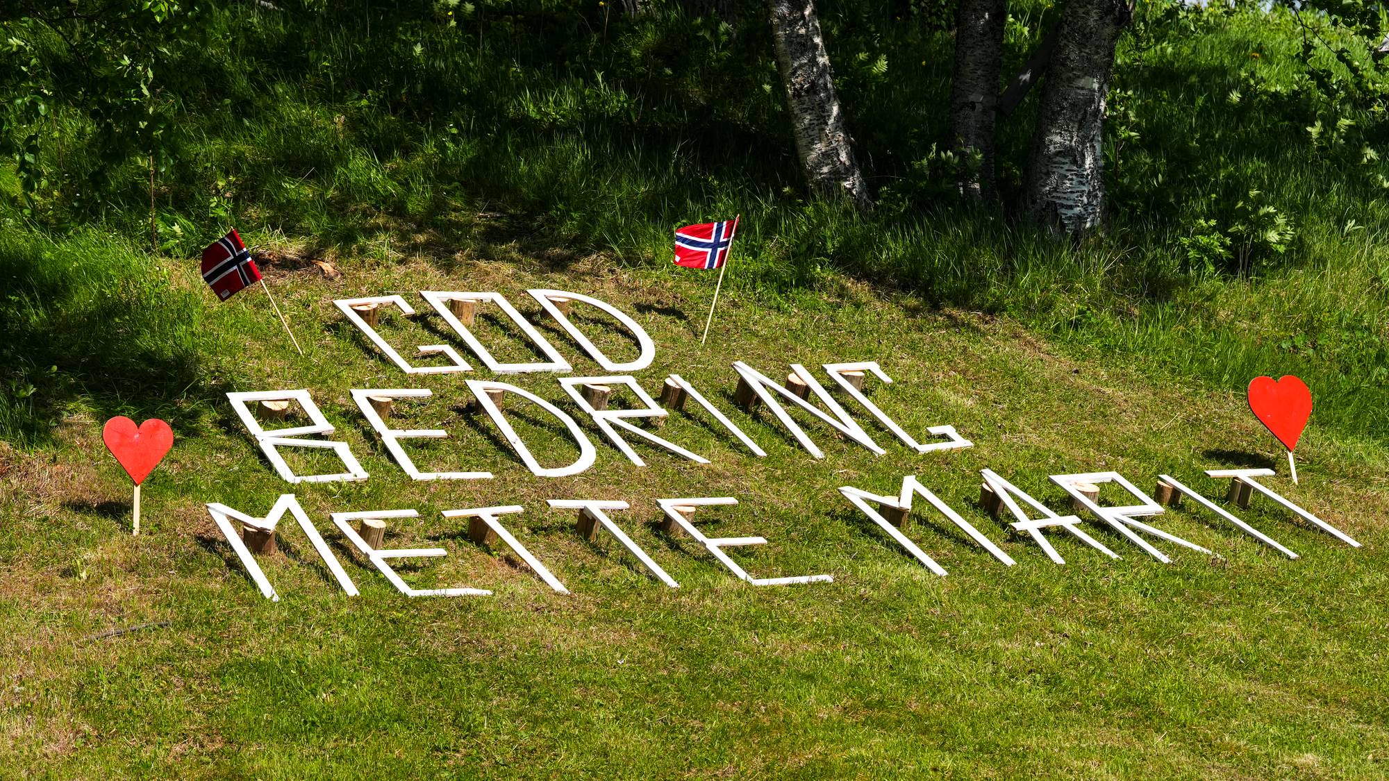 Teksten "GOD BEDRING METTE-MARIT" er stavet ut med hvite bokstaver på en gresskledd skråning. To norske flagg er plassert ved siden av teksten. To røde hjerter er også satt opp på pinner, et på hver side av budskapet. (Bildebeskrivelsen er laget av en KI-tjeneste)